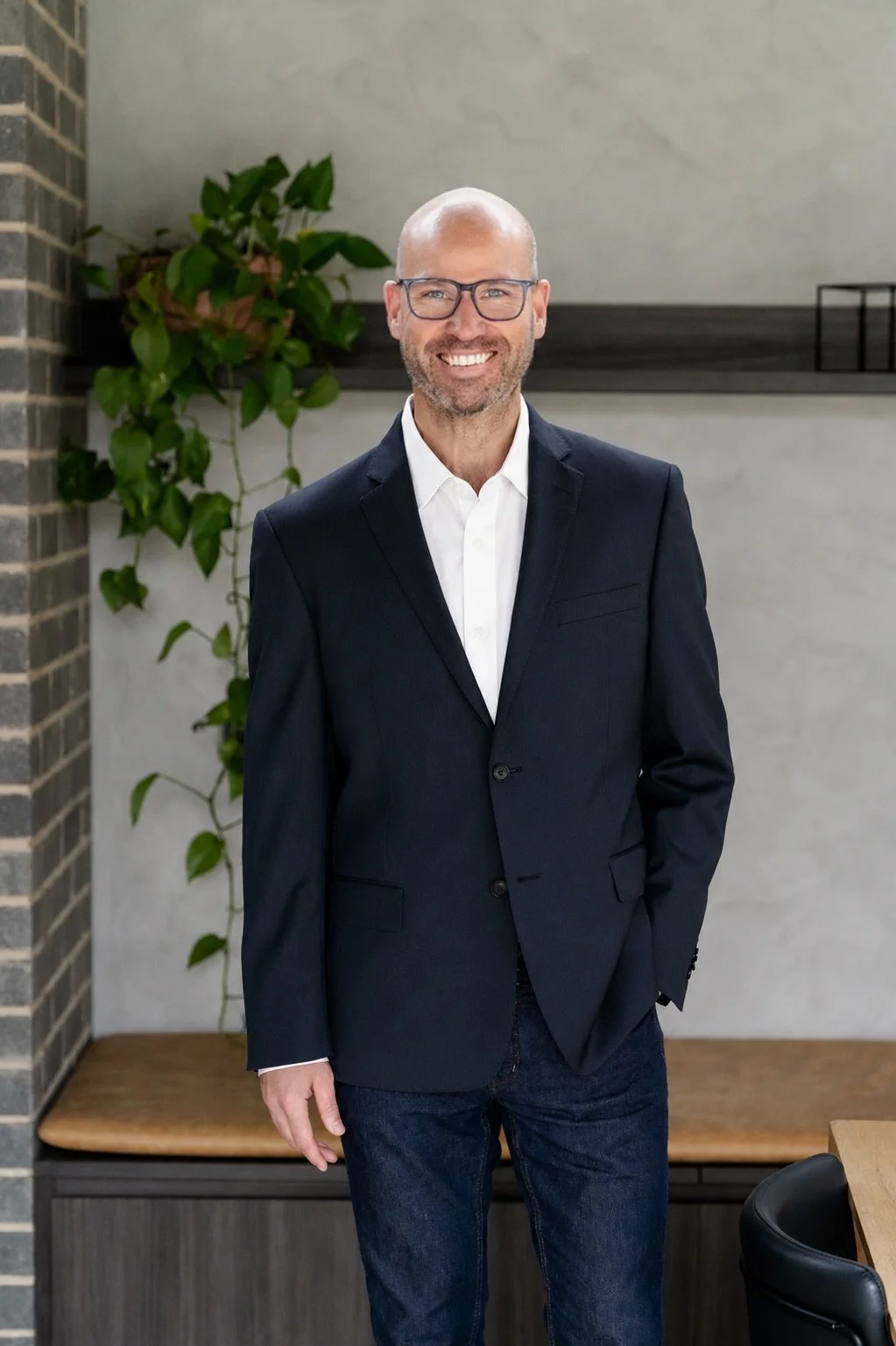 A man with glasses, a bald head, and a beard, wearing a dark blazer and jeans, smiling and standing in an indoor space with a plant and modern decor.
