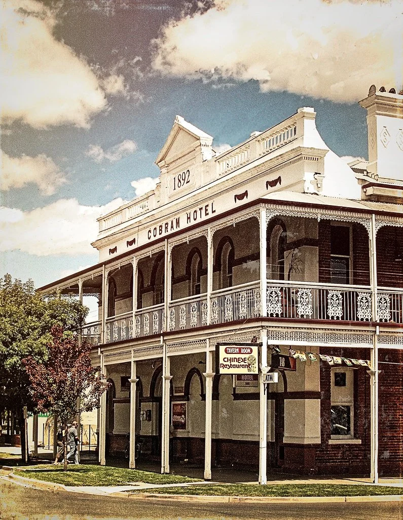 Historical two-story building labeled 'Cobbham Hotel' with white ornate balcony railings, a sign for a Chinese restaurant, and an ATM, under a partly cloudy sky.