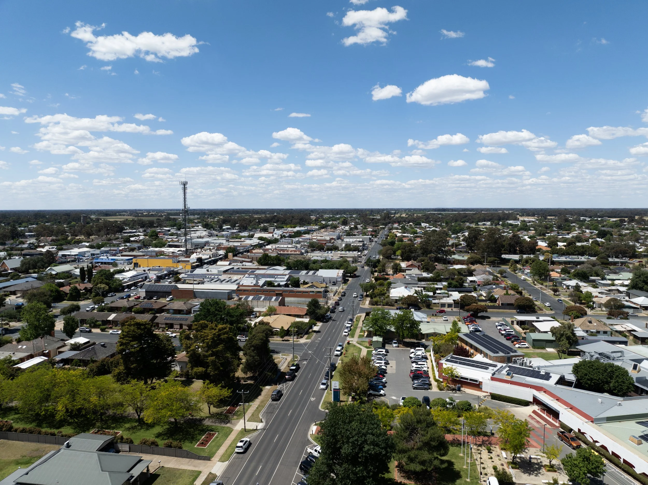 A cityscape view with a mix of residential and commercial buildings, a downtown area, and a wide street with vehicles. The sky is blue with scattered white clouds.