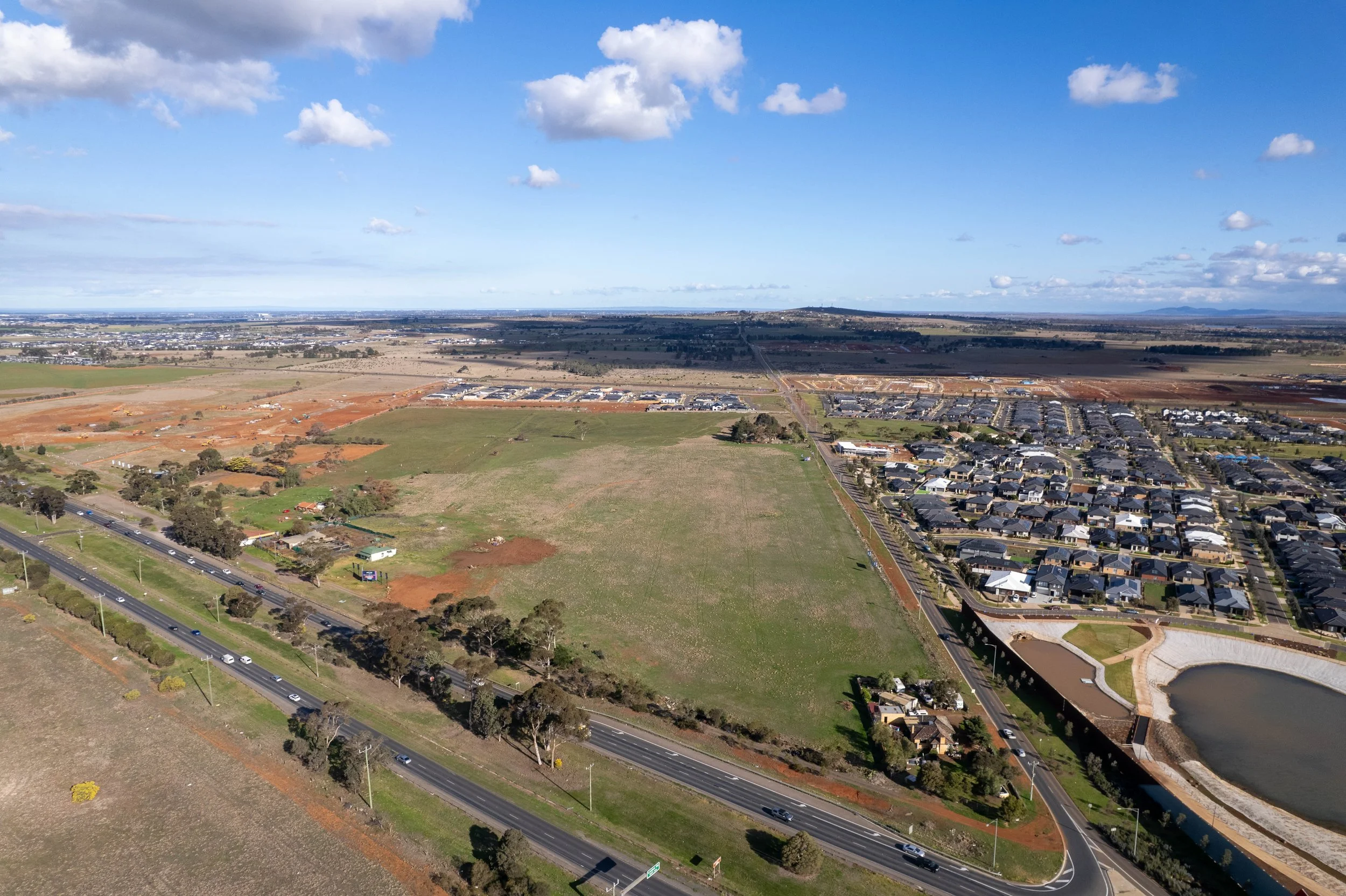 Aerial view of a suburban area with houses, roads, green fields, and open land under a partly cloudy sky.