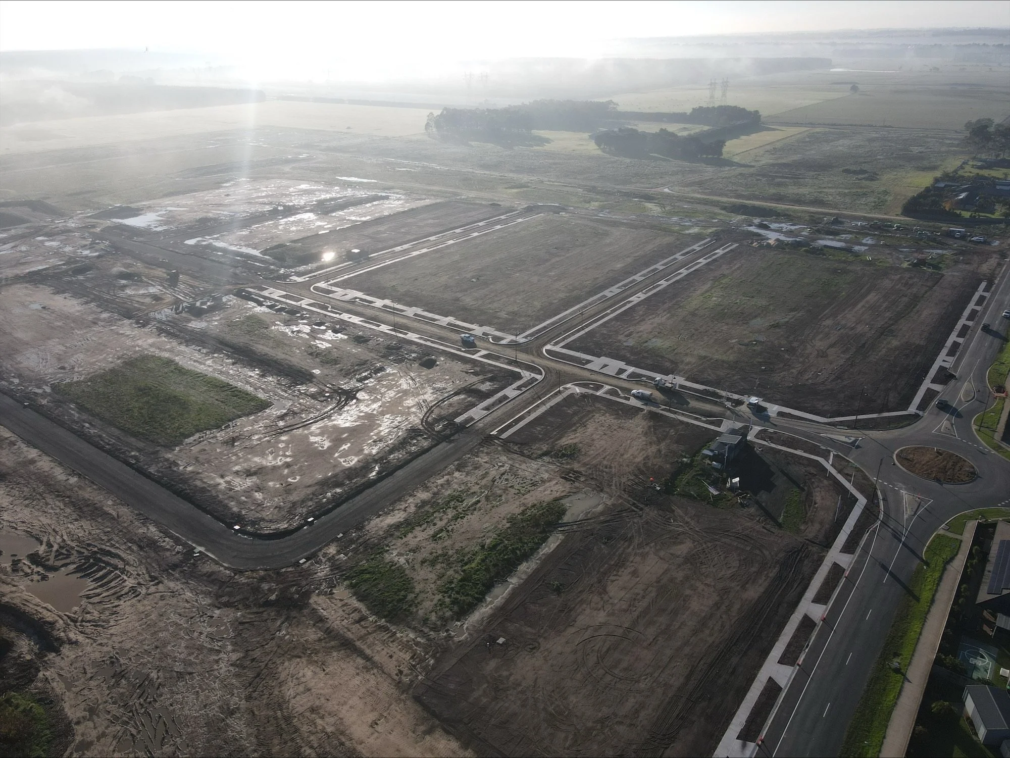 Aerial view of a construction site with dirt roads, gravel, and developing infrastructure, surrounded by fields and some trees.