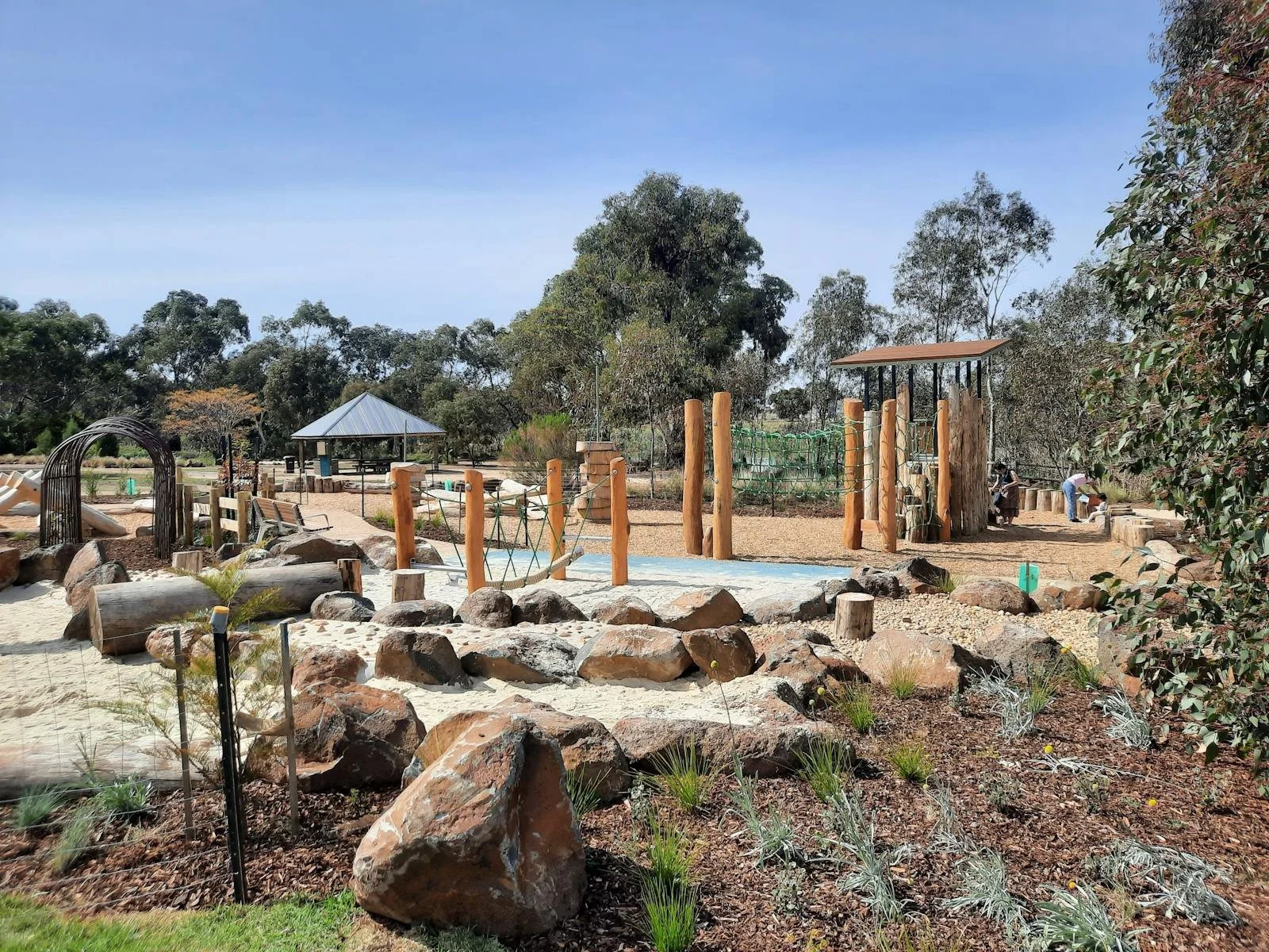 A playground with climbing structures, rocks, and trees in a park setting under a blue sky.