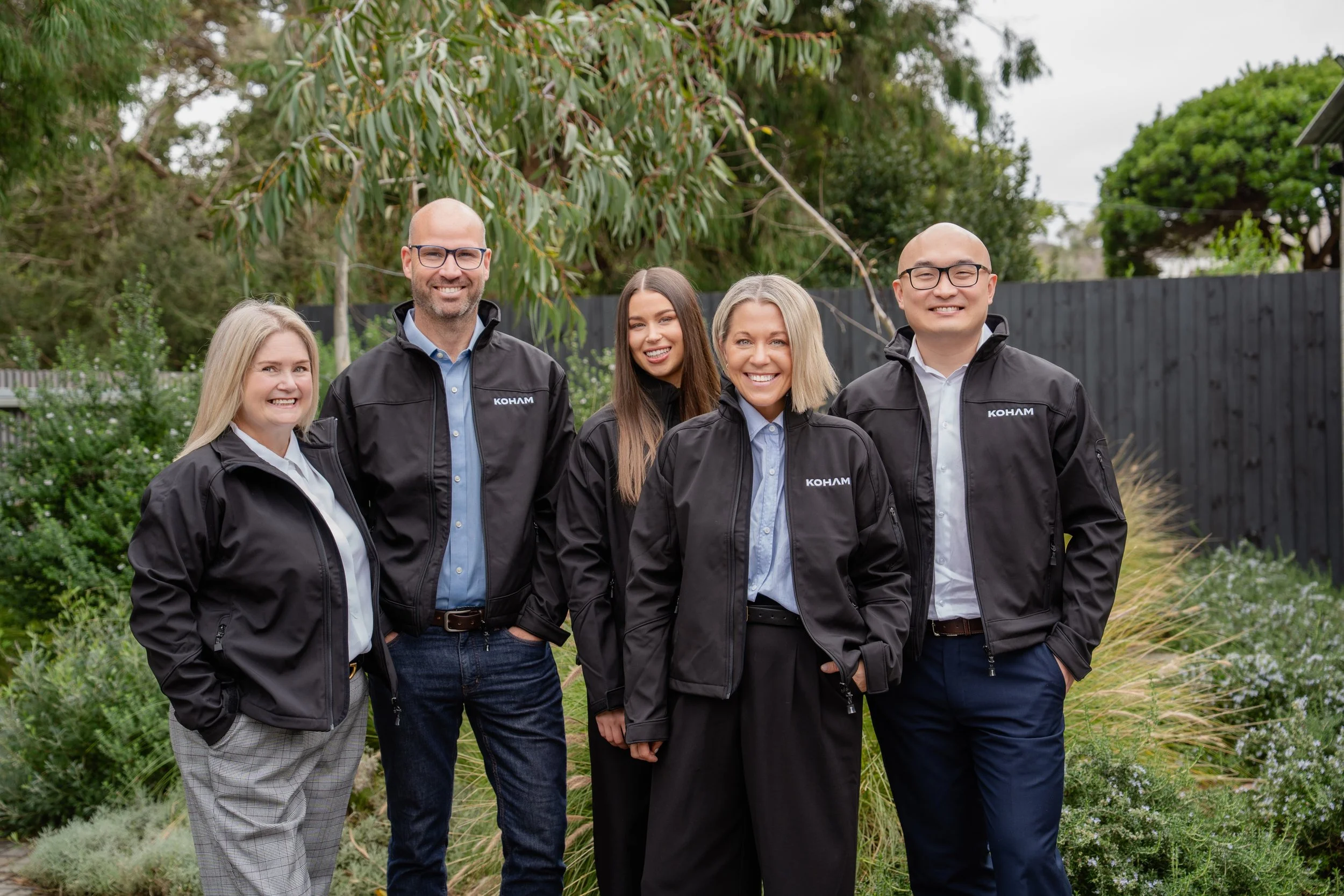 Group of five smiling diverse professionals standing outdoors in a garden area, wearing black jackets with the company's logo, with a background of trees and a wooden fence.