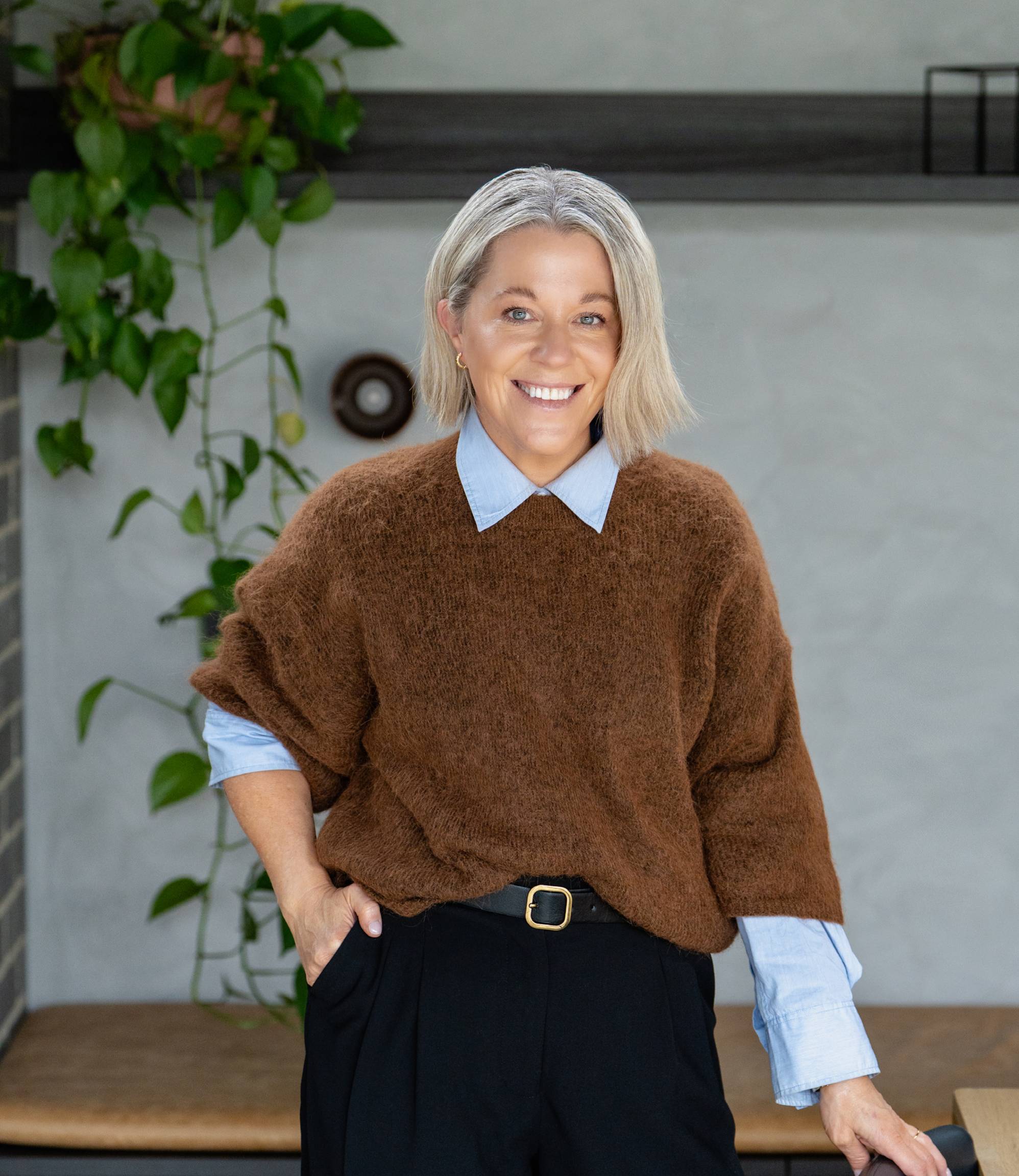 A woman with gray hair, wearing a brown sweater over a blue shirt, standing indoors in front of a gray wall with green plants and decorative objects behind her.