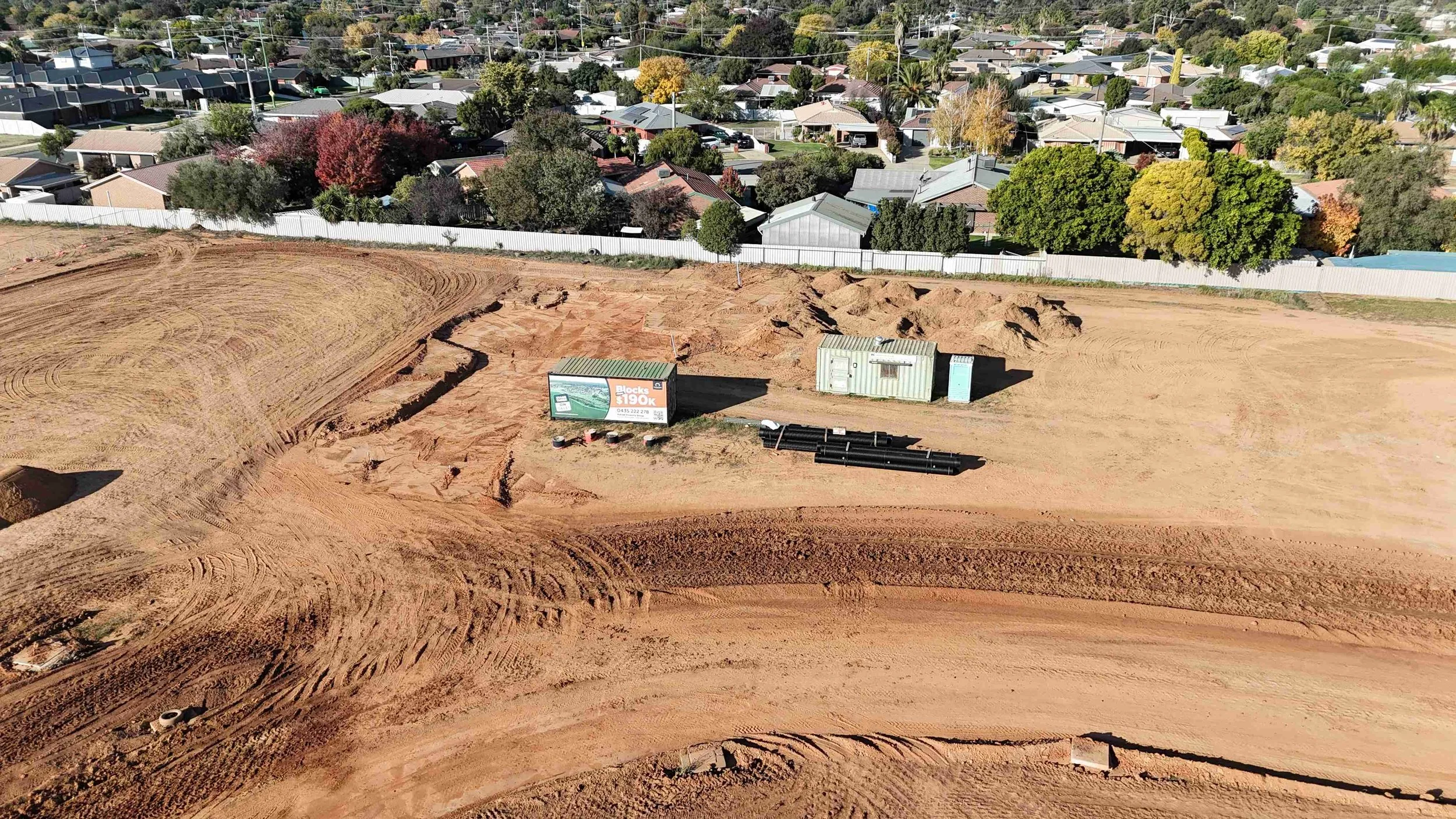 Construction site with dug-out earth, construction materials, and portable containers, surrounded by a residential neighborhood with houses, trees, and streets.