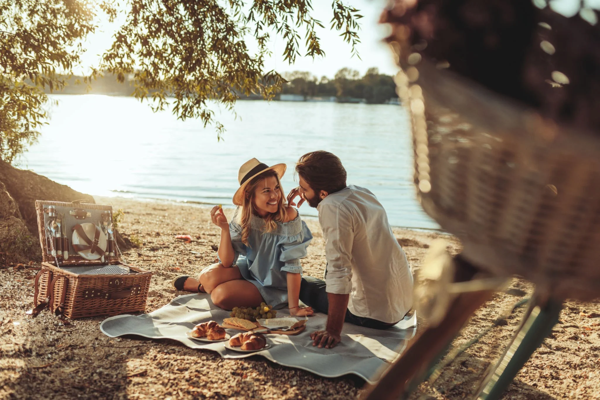 A couple having a picnic by the lakeside during sunset, with a picnic basket and food on a blanket, surrounded by trees.