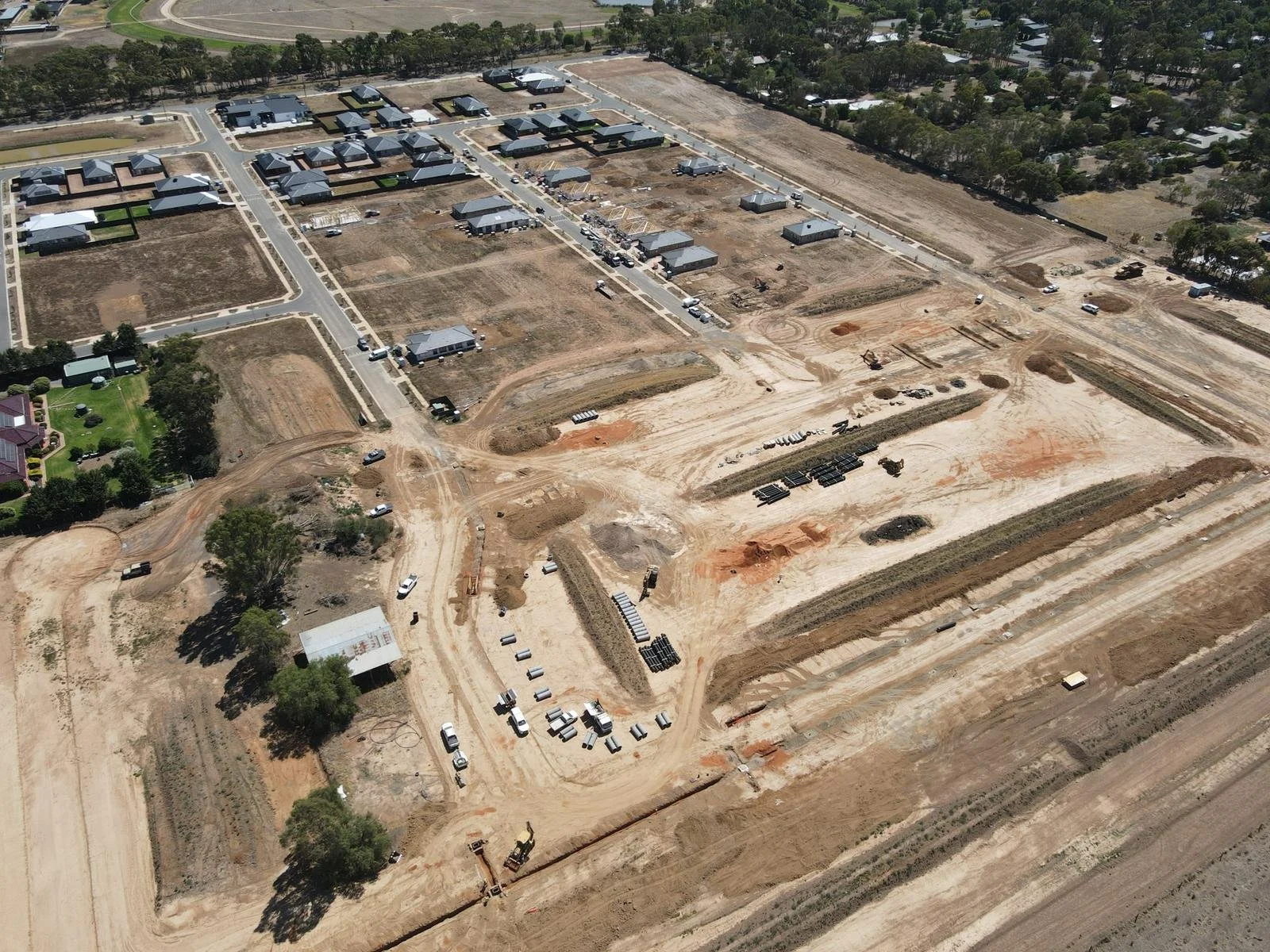 Aerial view of a construction site with cleared land, construction materials, and partially built structures, adjacent to a neighborhood with houses and trees.