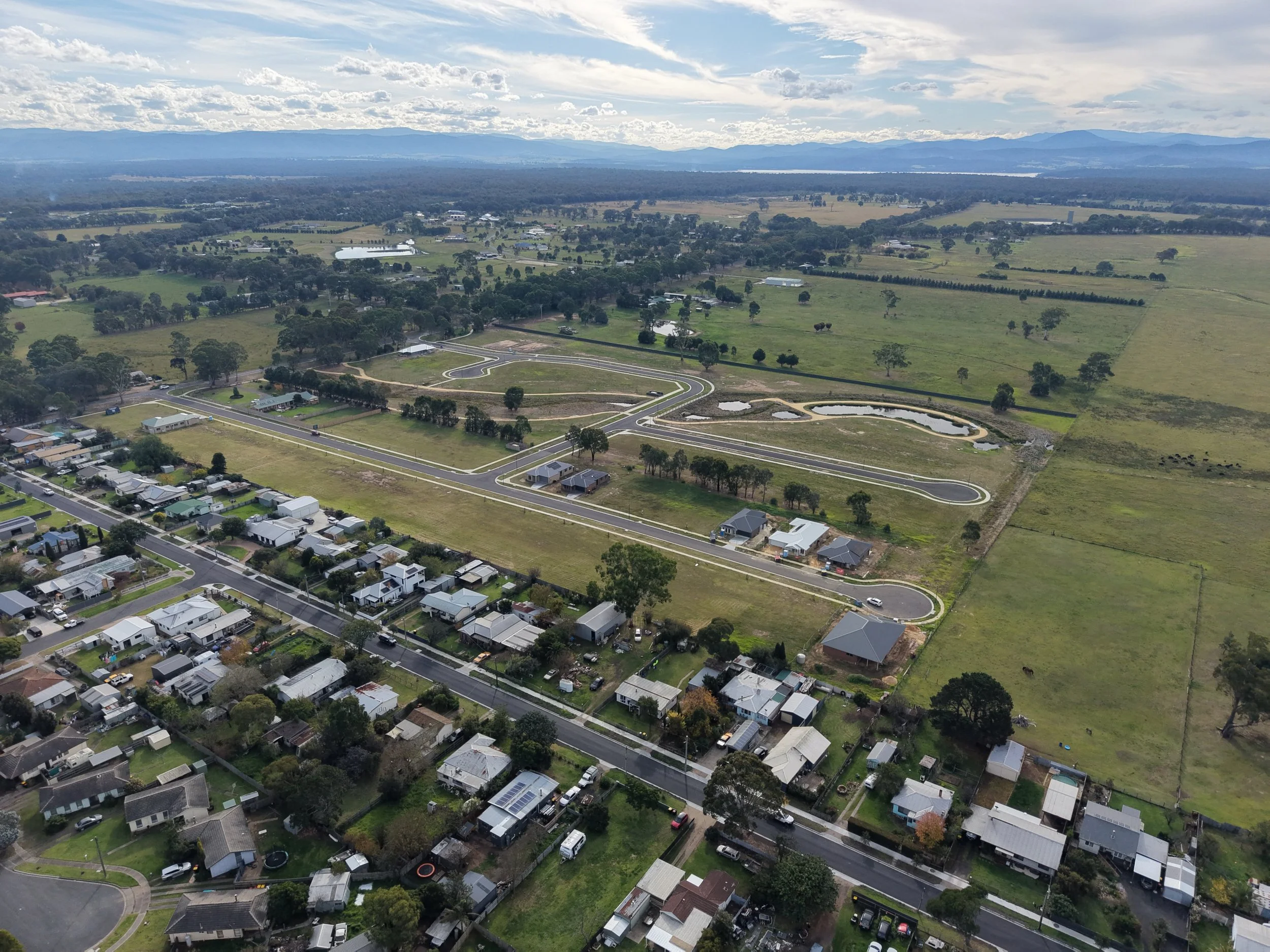 Aerial view of a suburban neighborhood with houses, streets, trees, a winding road, small ponds, and expansive green fields. In the background, there are mountains under a partly cloudy sky.
