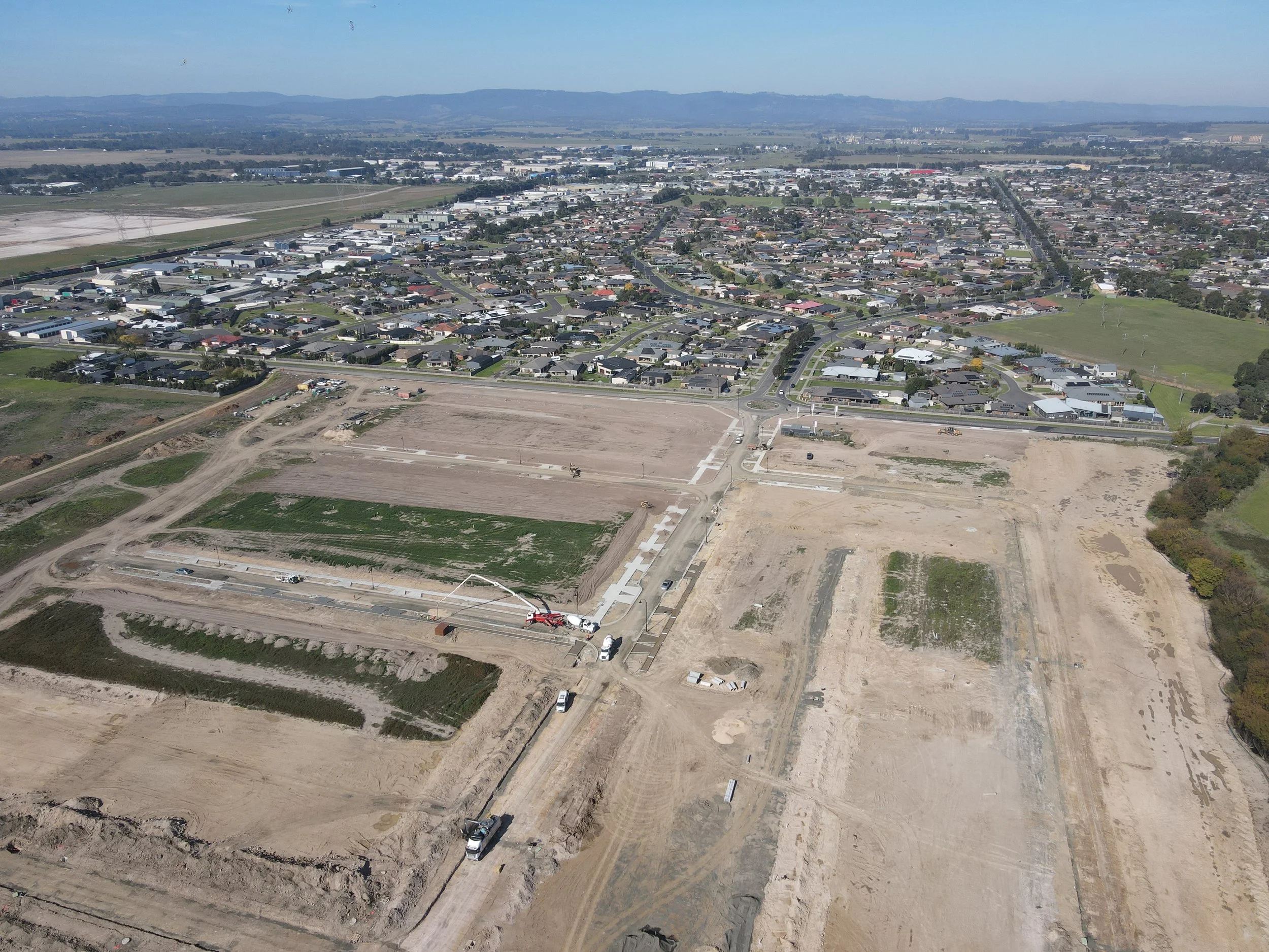 Aerial view of a construction site in a suburban area with roads, houses, and undeveloped land, mountains in the background.