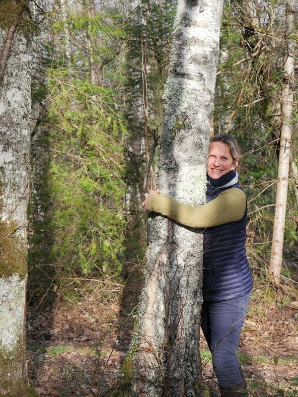 Une femme souriante en pull beige et veste noire, donnant un câlin à un arbre dans une forêt.