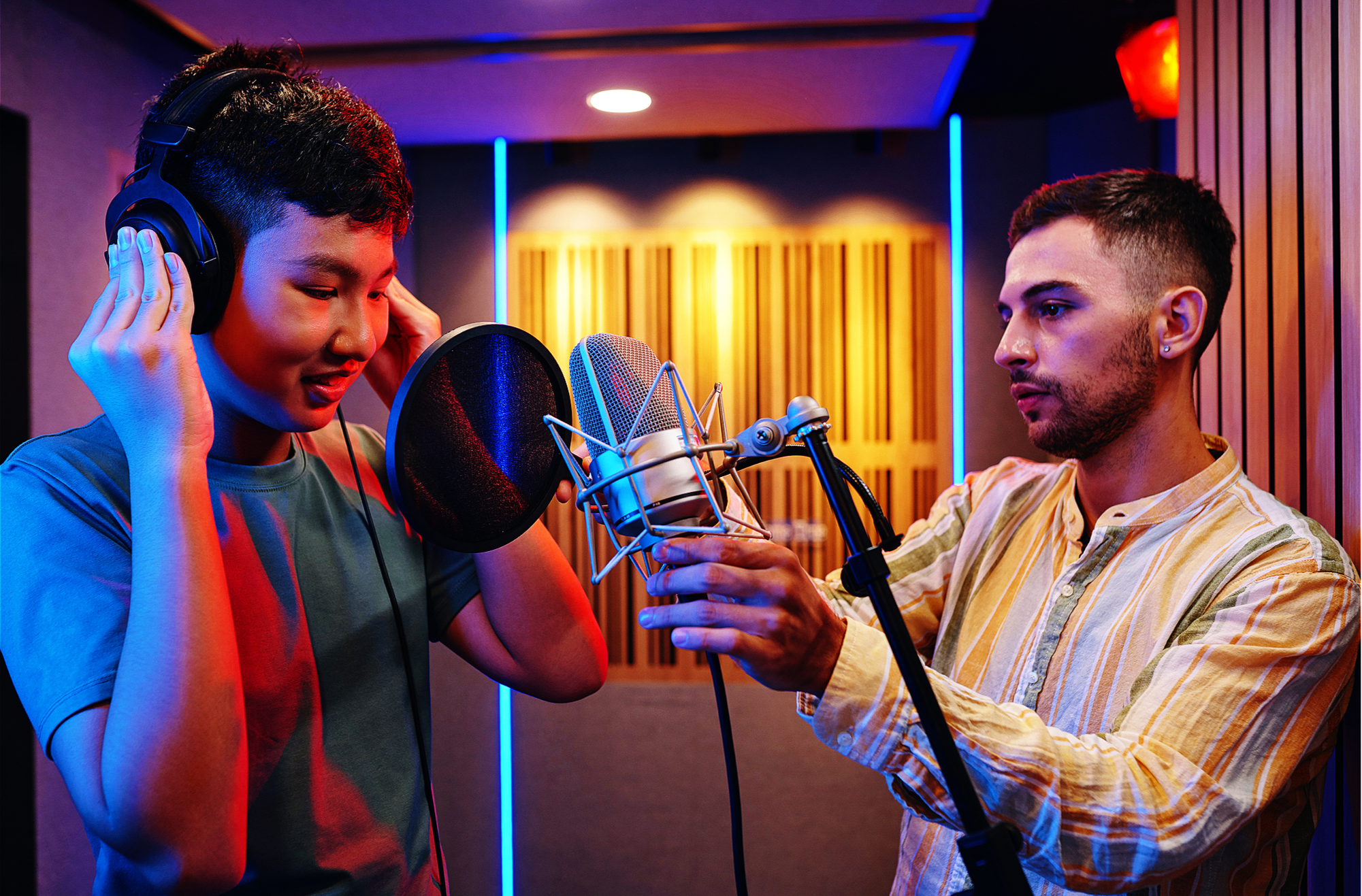 Two men in a recording studio, one wearing headphones and speaking into a microphone, the other adjusting the microphone, with acoustic panels and colorful lighting in the background.