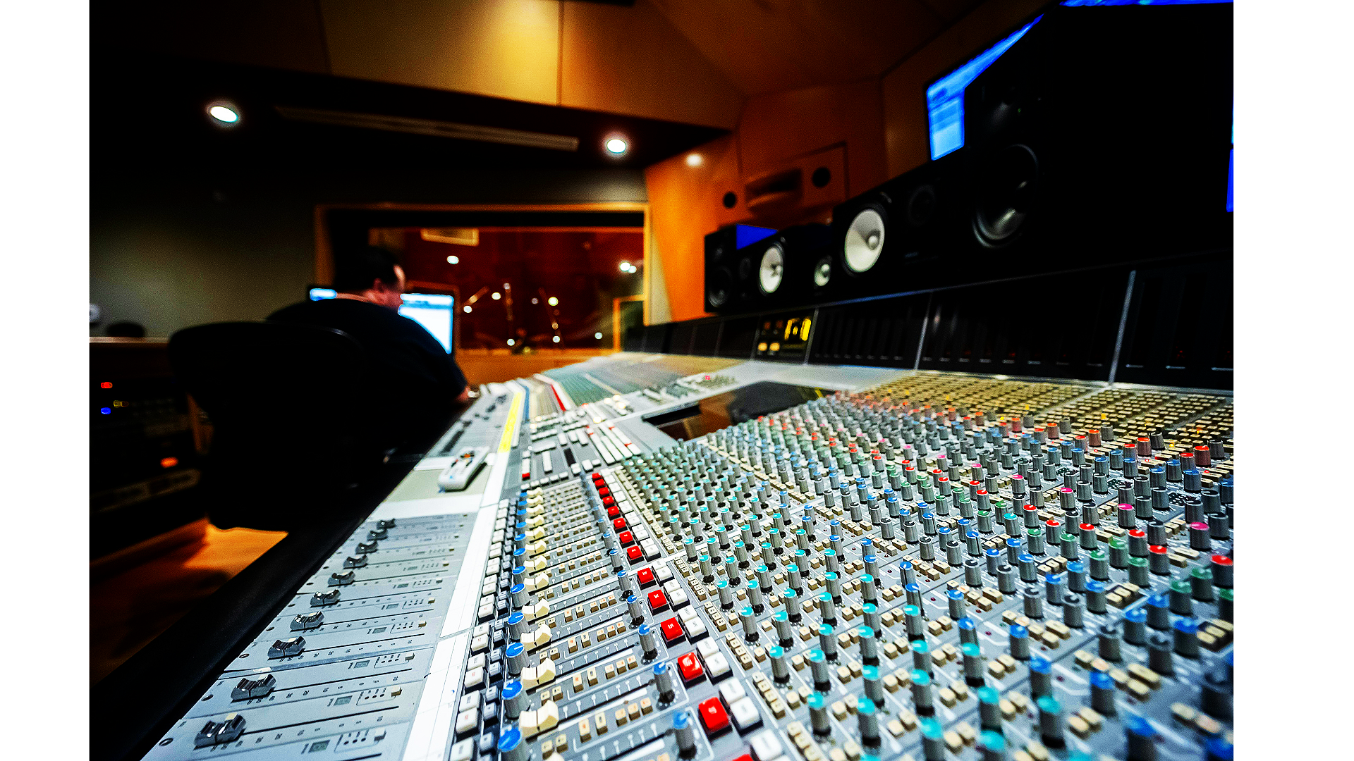 Close-up of audio mixing console with colorful faders and knobs in a recording studio, with a person working at a computer in the background.