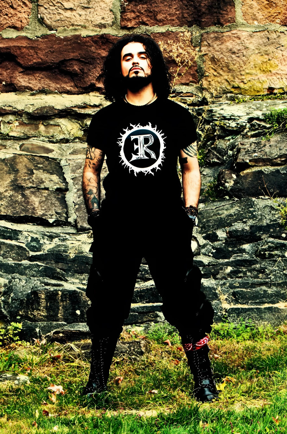A man with long curly hair and tattoos on his arms standing outdoors in front of a stone wall, wearing a black t-shirt with a large logo and black pants.