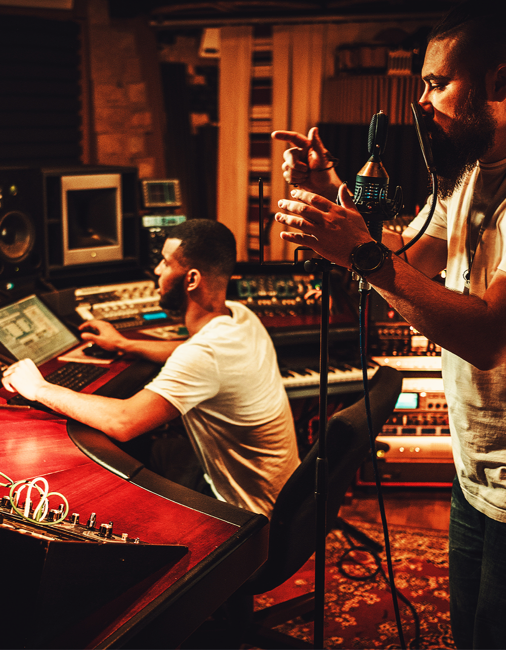 Two men in a music recording studio, one at a mixing console working on audio, the other standing next to a microphone speaking or singing.