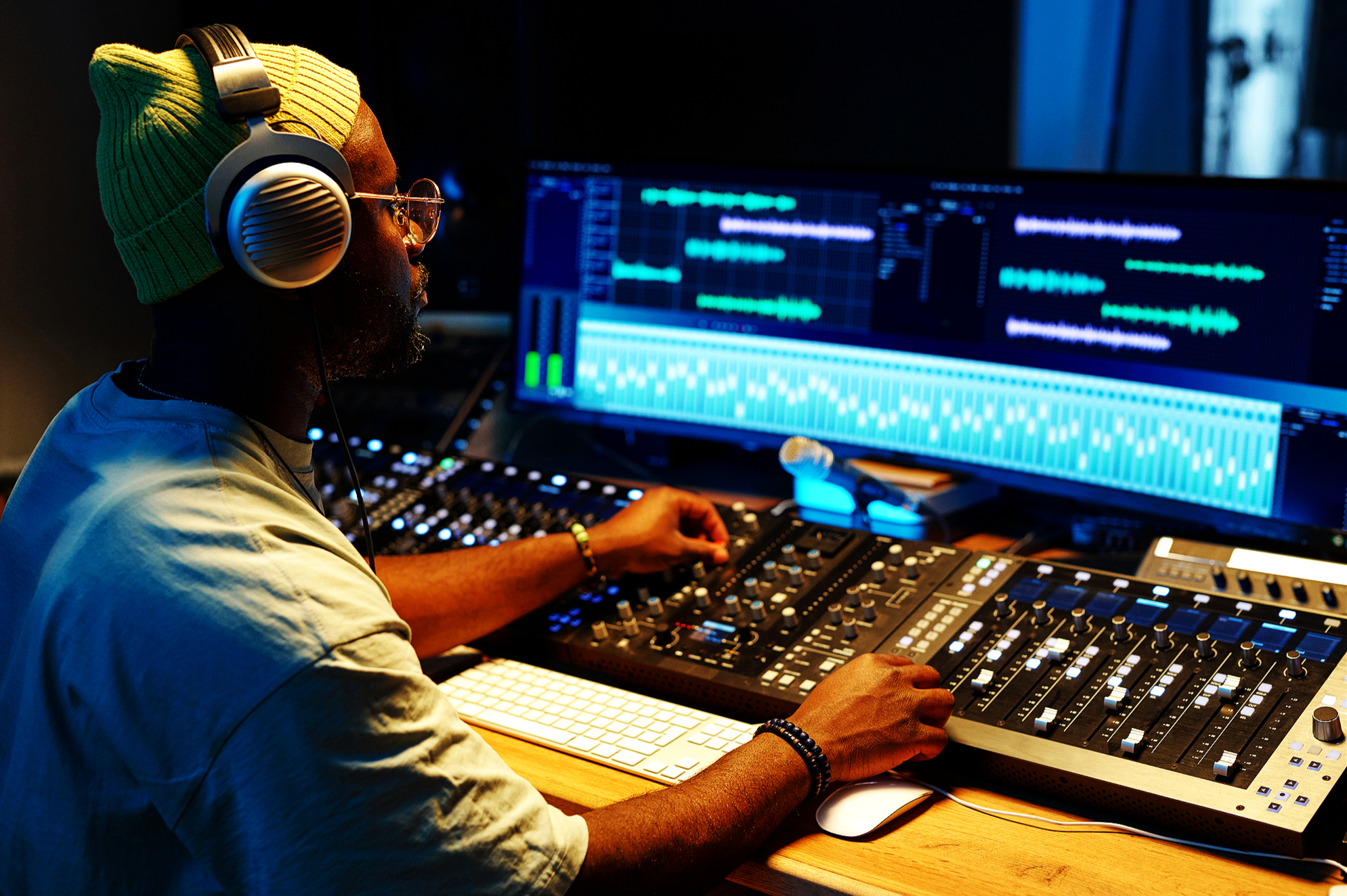 A man in a yellow beanie and glasses, wearing headphones, working at a music production station with a mixing console, computer monitors displaying audio editing software.