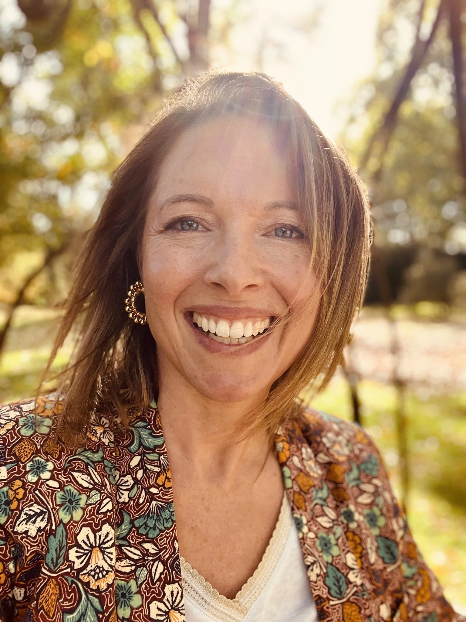 A smiling woman with shoulder-length light brown hair outdoors in sunlight, wearing a floral-patterned jacket, gold hoop earrings, and a white top, with trees and sunlight in the background.