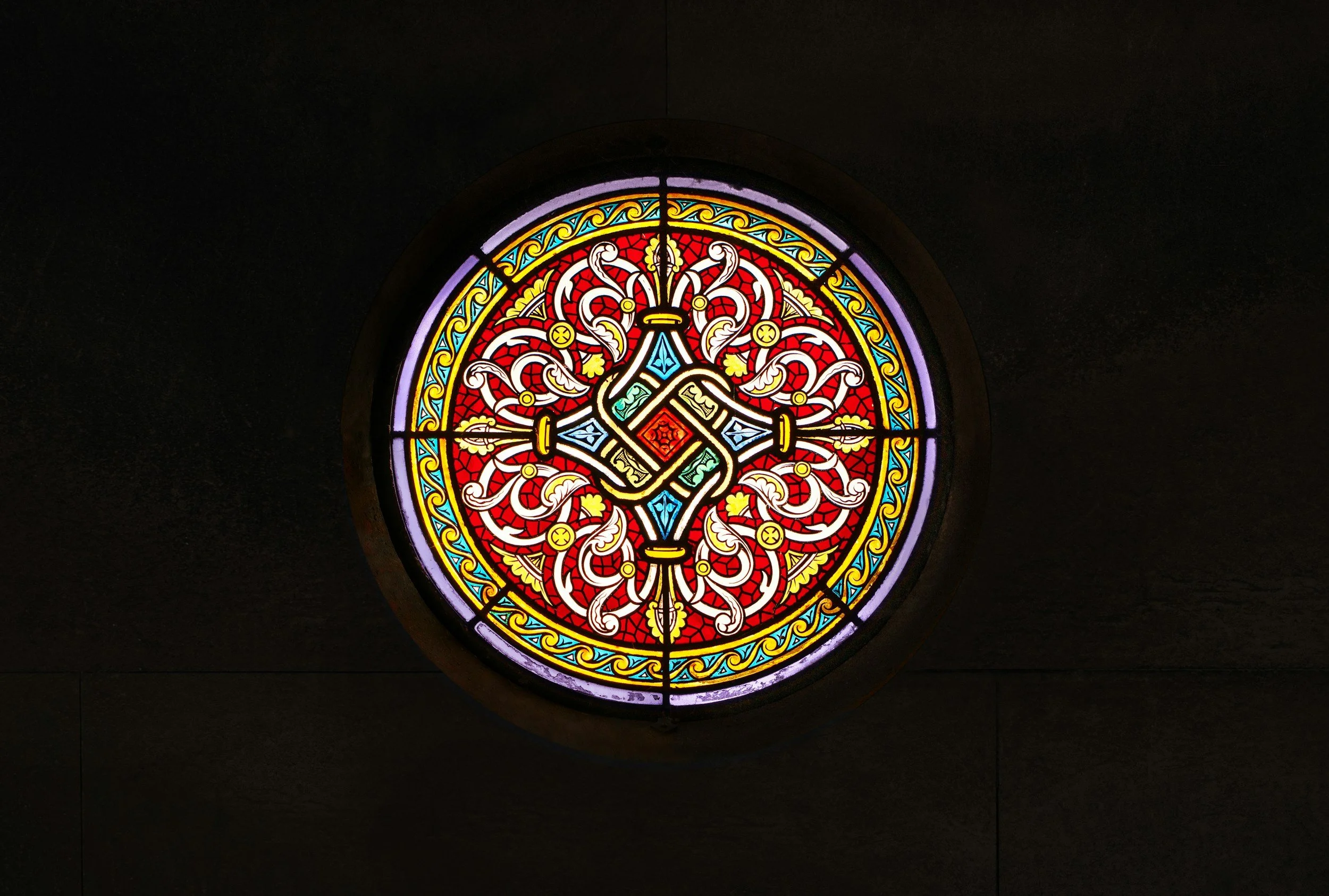 Colorful stained glass window with intricate geometric and floral designs, predominantly in red, yellow, blue, and green.