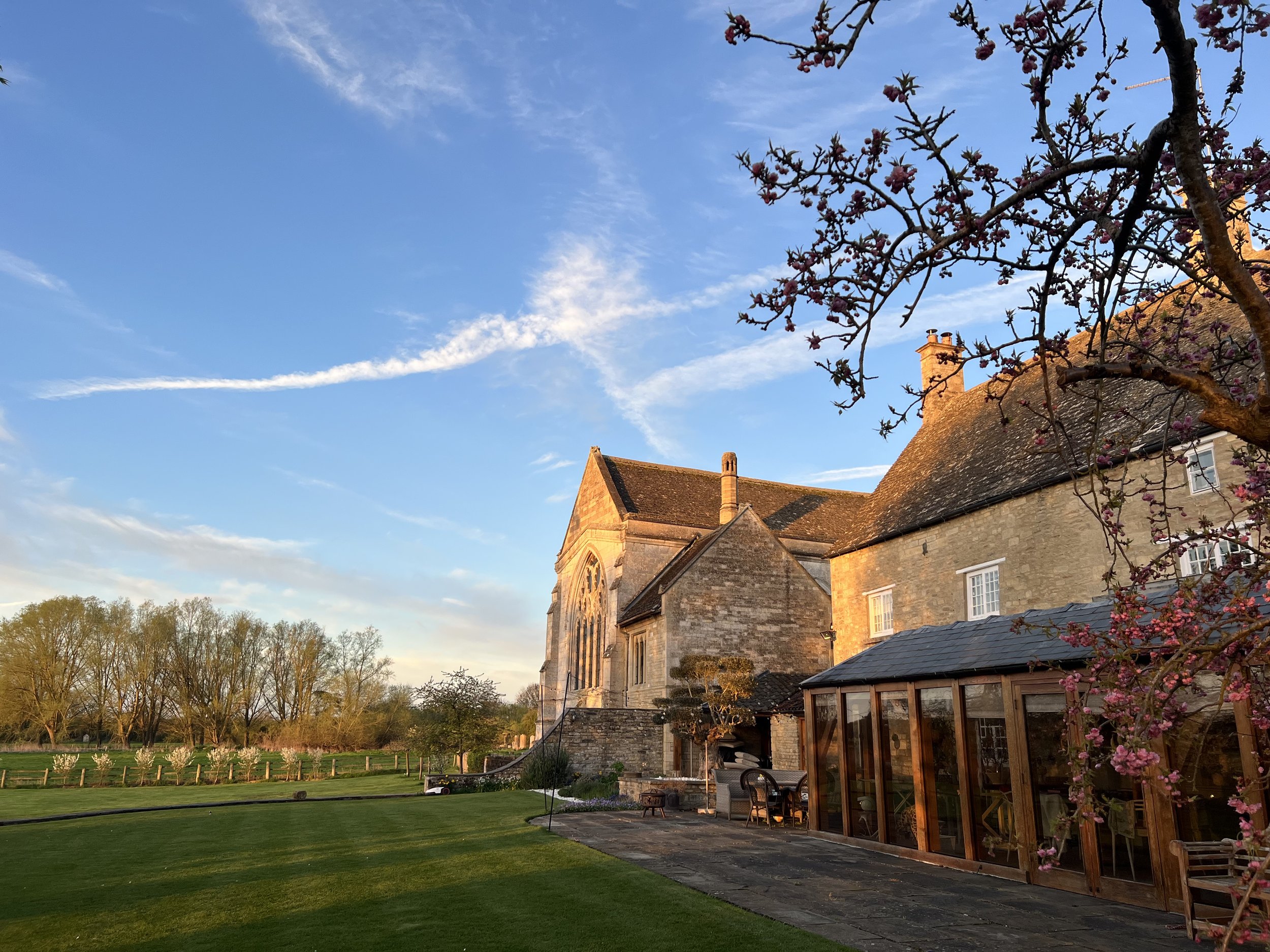 A historic stone building with a large windowed conservatory, a stone pathway, pink flowering trees, a lawn, and a blue sky with some clouds and contrails.