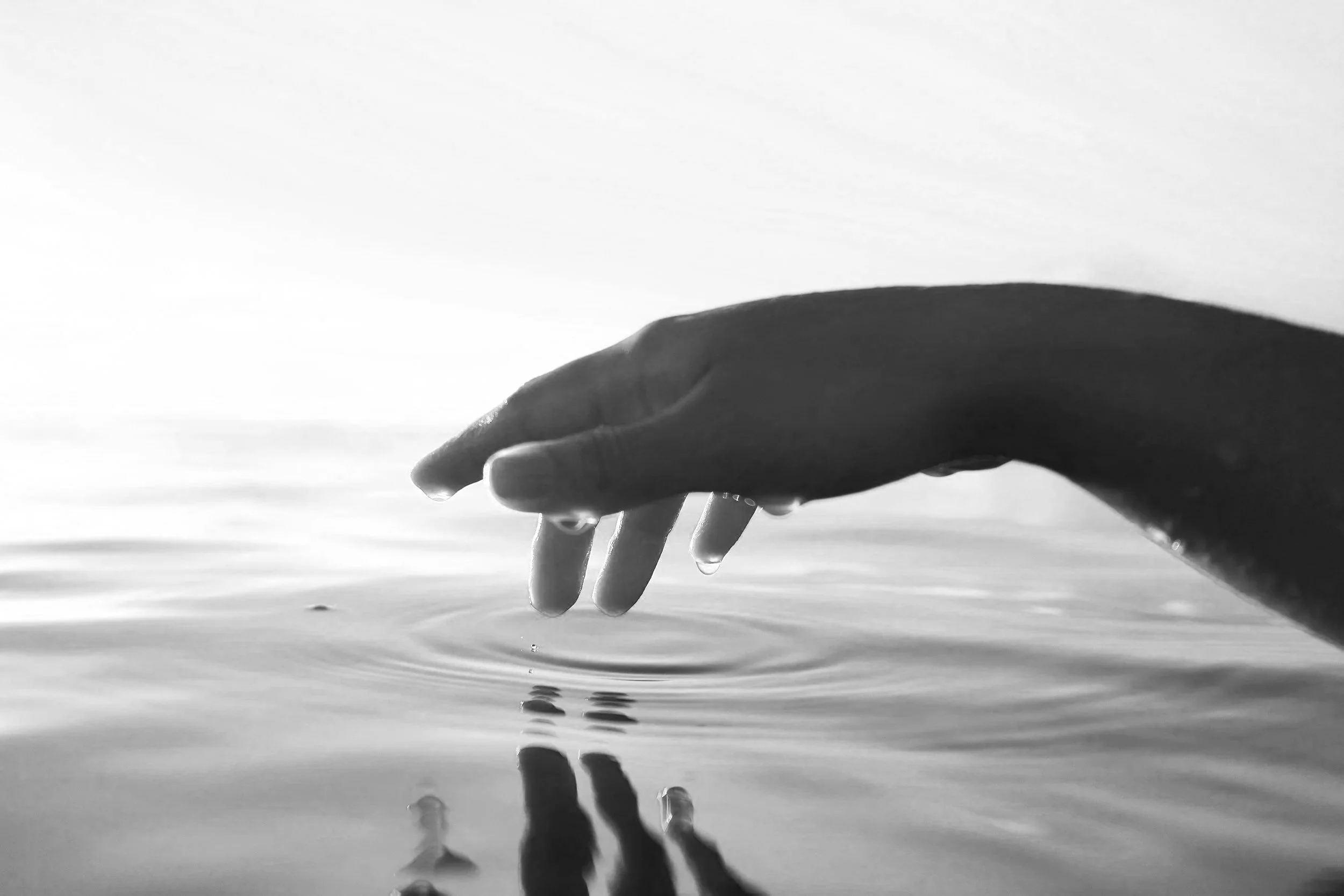 A person's hand touching the surface of calm water, creating ripples, in black and white.