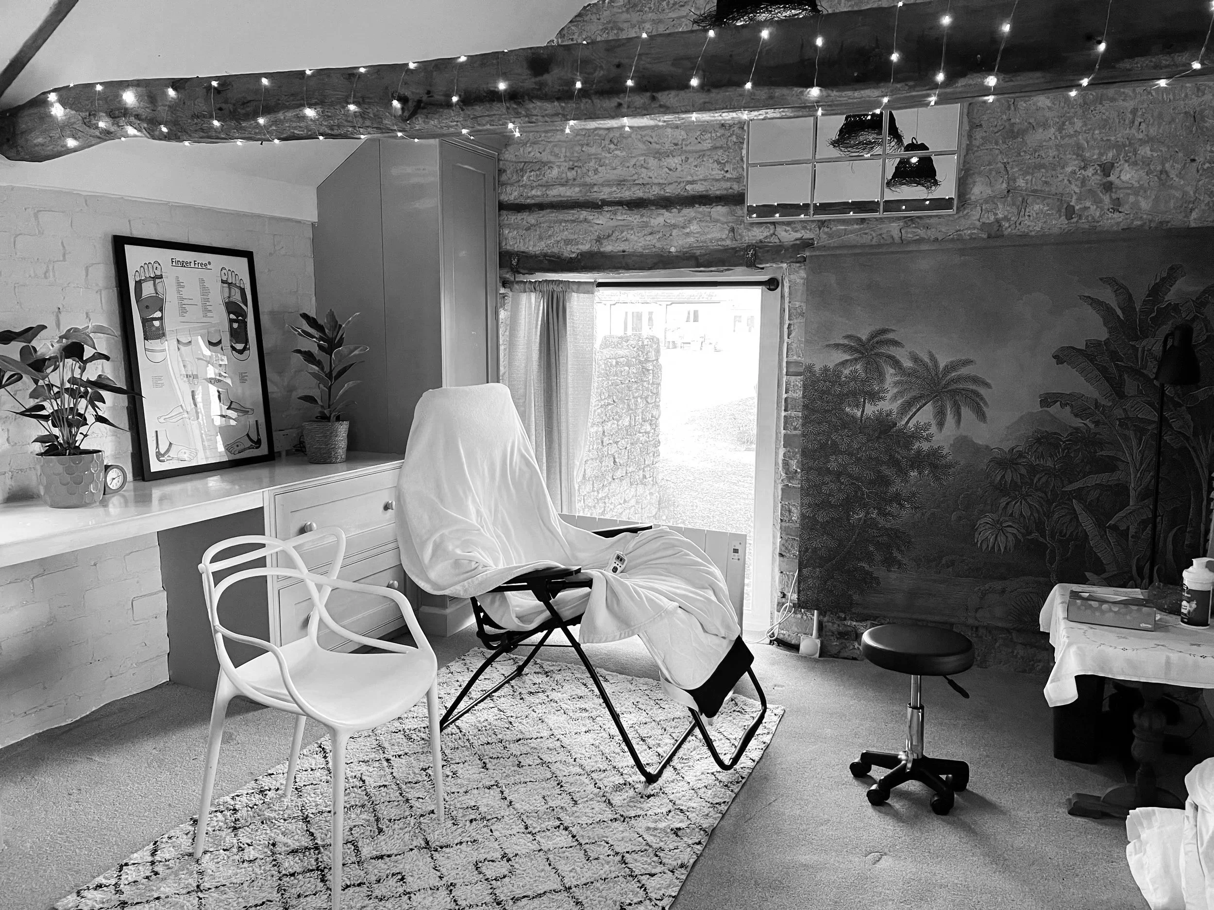 A black and white photo of a room with a medical examination chair covered with a sheet, a white modern chair, a small table with supplies, potted plants, framed artwork, a wall mural of trees, a window with a curtain, string lights on a wooden beam, and a swivel stool.