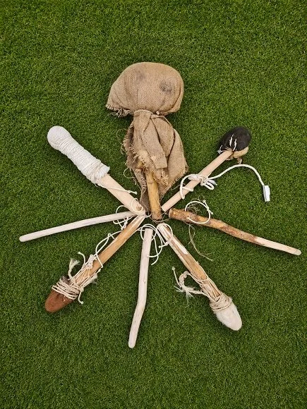 A stick figure doll made of sticks, cloth, and various objects, lying on a grassy surface.