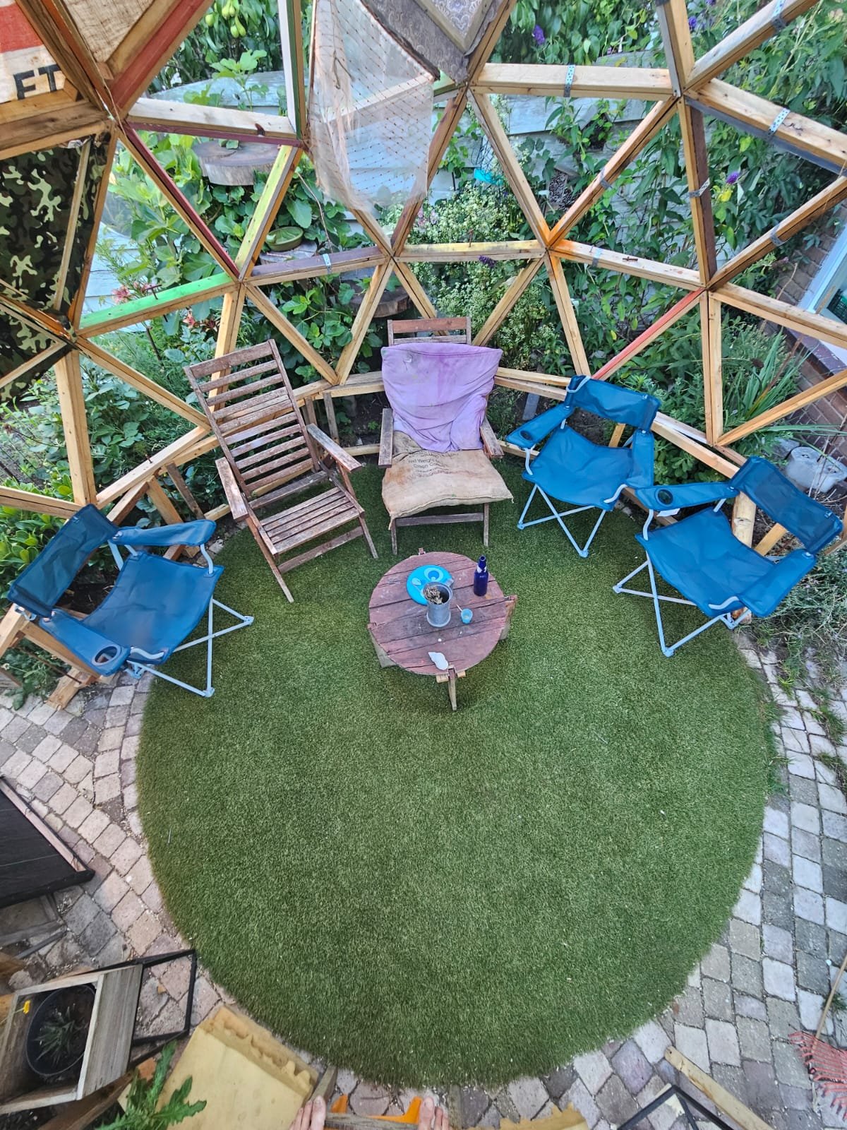 A small outdoor seating area with a green artificial grass rug, four blue folding chairs, and two wooden chairs, one with a purple cloth draped over it. There is a small round wooden table in the center with various items on it, surrounded by a supporting wooden geodesic dome structure with greenery visible through the gaps.