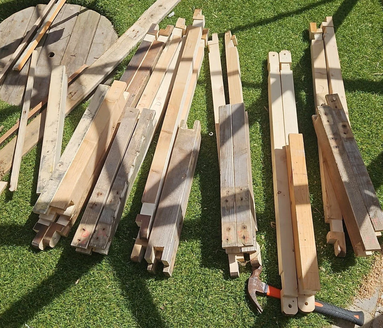 Various wooden planks and pieces laid out on grass for construction, with tools including a hammer and a saw nearby.