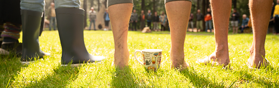 Mensen staan op gras, met een kopje in het centrum en anderen om hen heen, in een park met veel mensen op de achtergrond.
