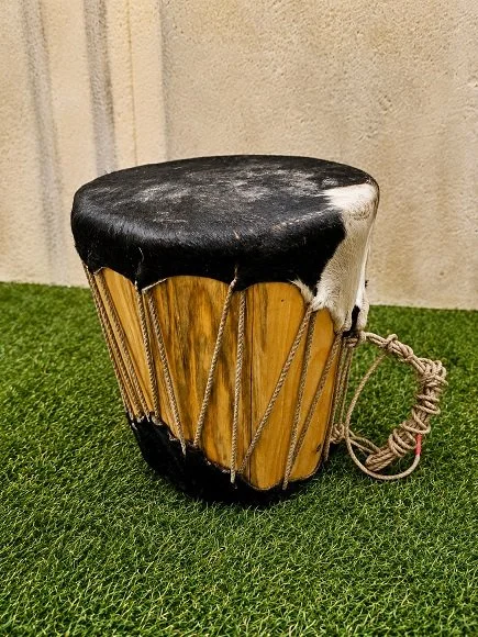 Traditional hand drum with a wooden body, black animal hide top, and tied strings around the body, placed on green grass.