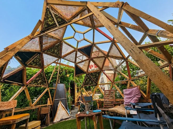 Interior of a small, partially open wooden geodesic dome with furniture including chairs, cushions, and a small table, surrounded by green plants and blue sky.