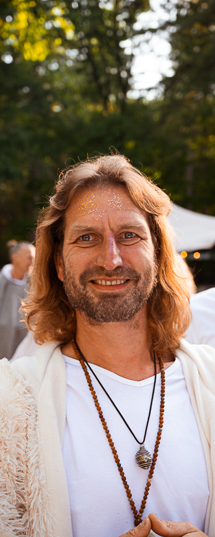 Man met lange haren, glimlach, gele glitter op voorhoofd, buiten op een zonnige dag.