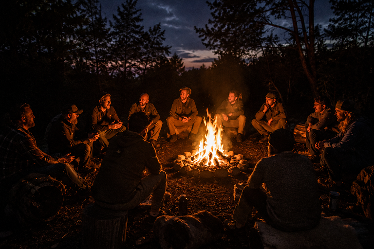 Een groep mensen zit in een cirkel rond een kampvuur in een bos in de avond. Ze lijken te genieten en hebben een ontspannen sfeer.