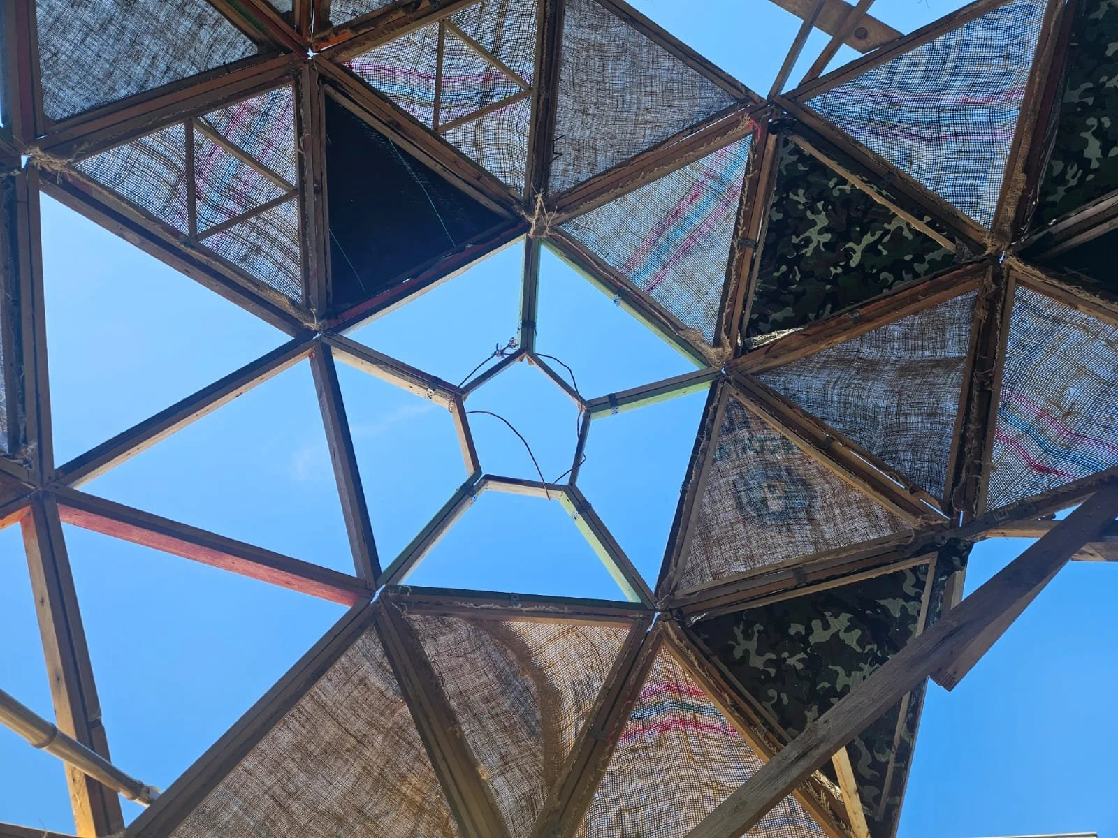 View of a geometric wooden and fabric structure viewed from below with the sky visible through the open center.