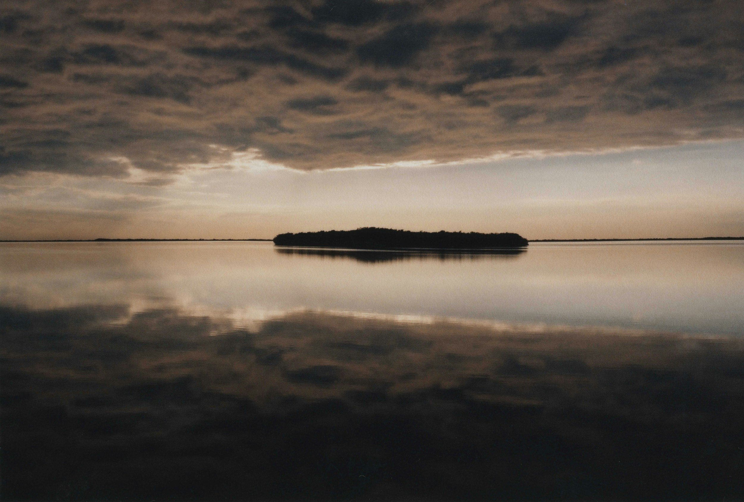 A body of water reflecting dark, cloudy sky during sunset or sunrise with a silhouette of land or island in the distance.