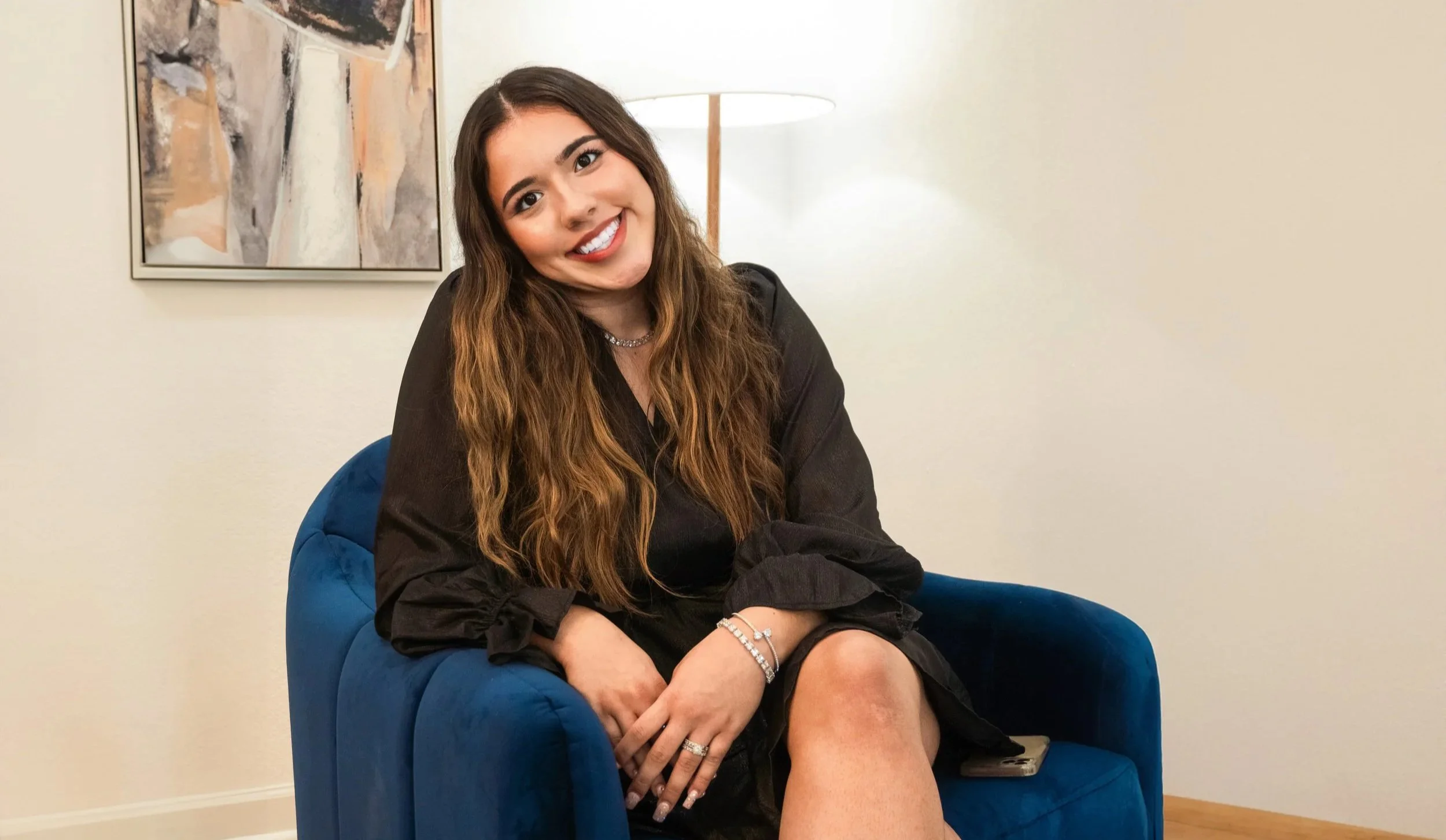 A woman with long wavy brown hair smiling and sitting on a blue velvet armchair in a living room with a framed abstract painting on the wall and a tall standing lamp in the background.