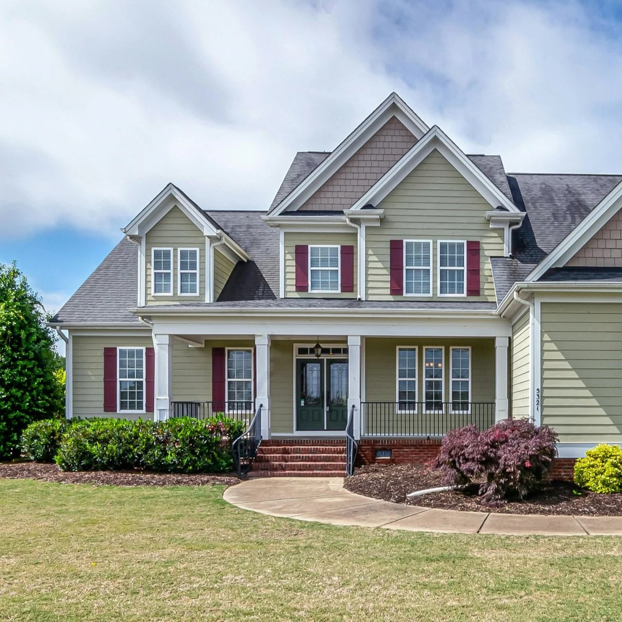 A large yellow house with white trim, red shutters, and a front porch, surrounded by green shrubs and a small tree.