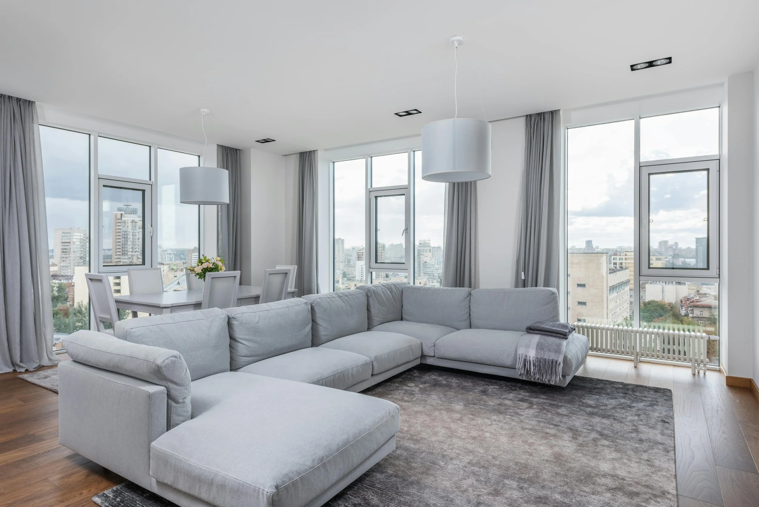 Modern living room with large windows, a light gray sectional sofa, a dark gray area rug, and a dining area with a white table and chairs, city view outside.