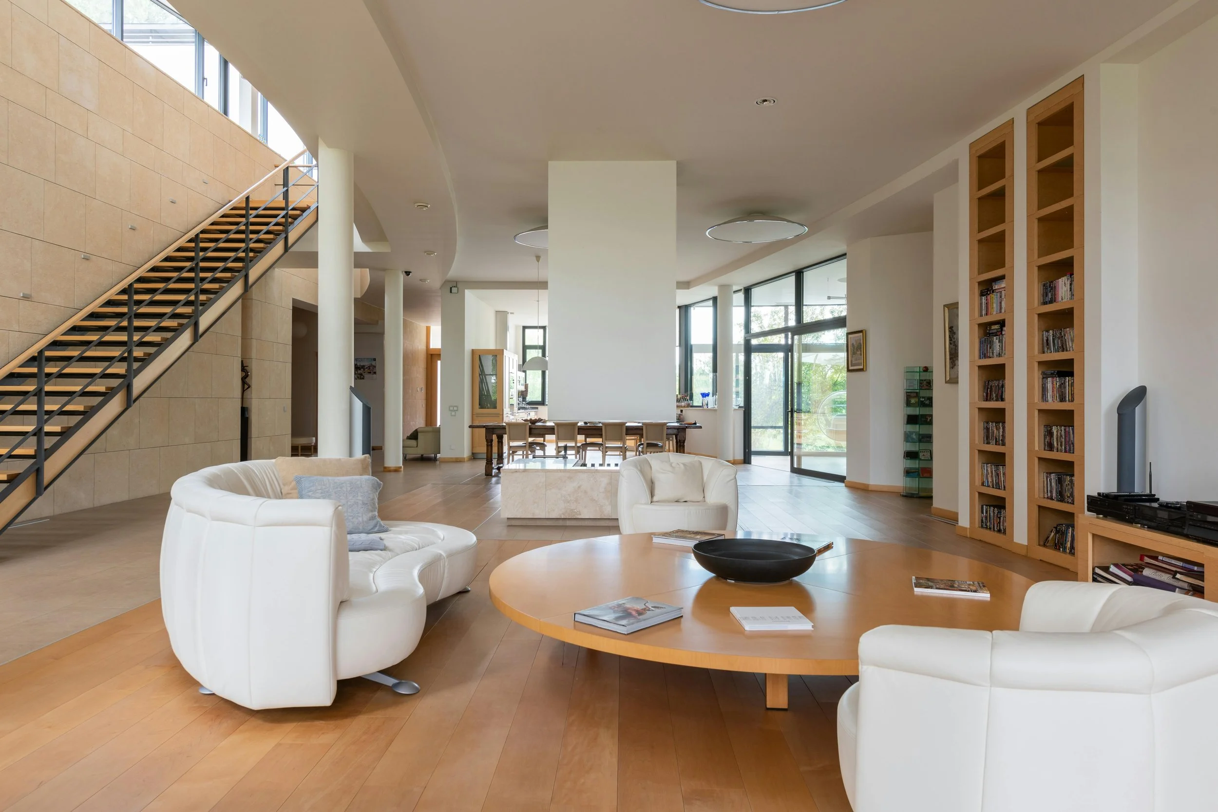 Spacious modern living room with wooden floors, white leather chairs, a round wooden coffee table, a large built-in bookshelf filled with DVDs, and a staircase with black railing leading upstairs. Large windows let in natural light.