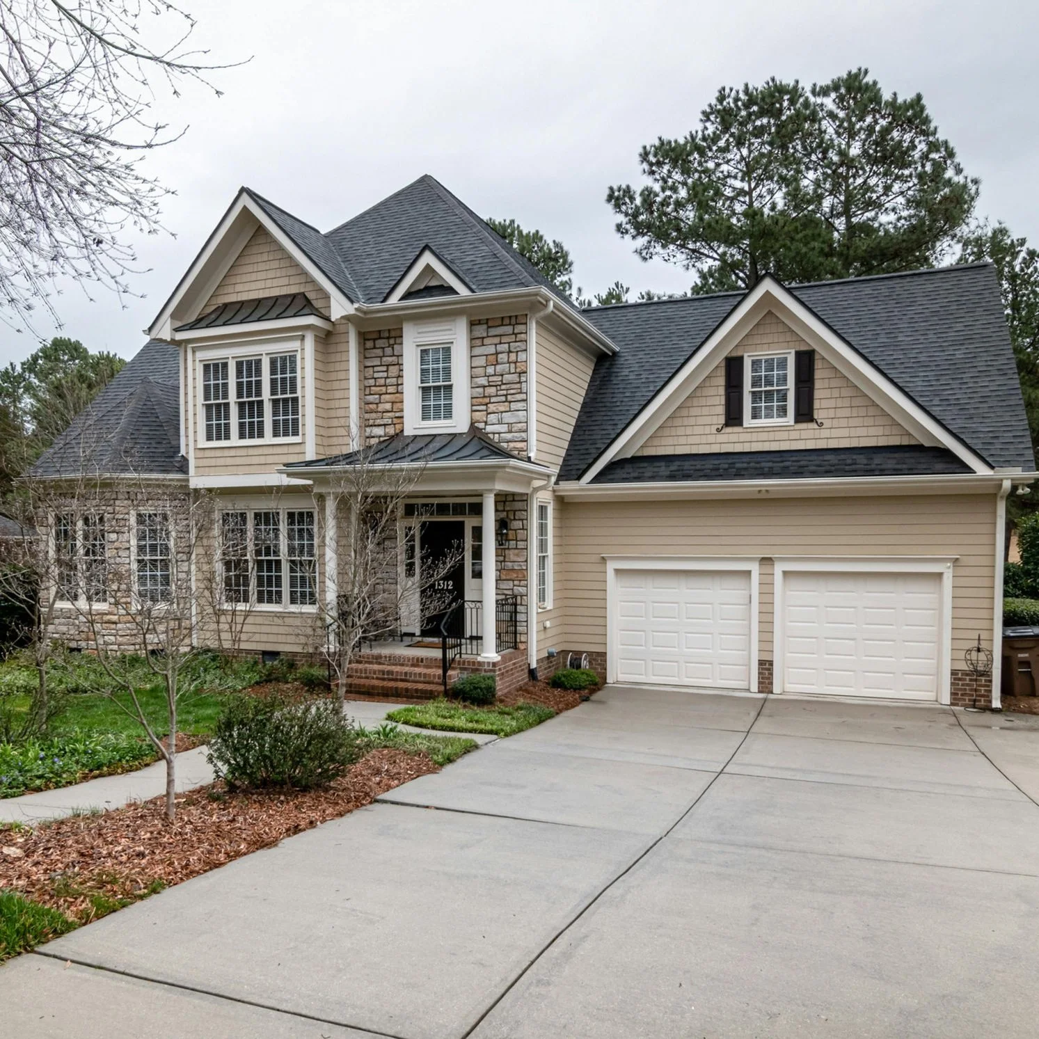 A two-story house with beige siding, brick accents, multiple gables, and black shutters, surrounded by a manicured lawn and leafless trees.