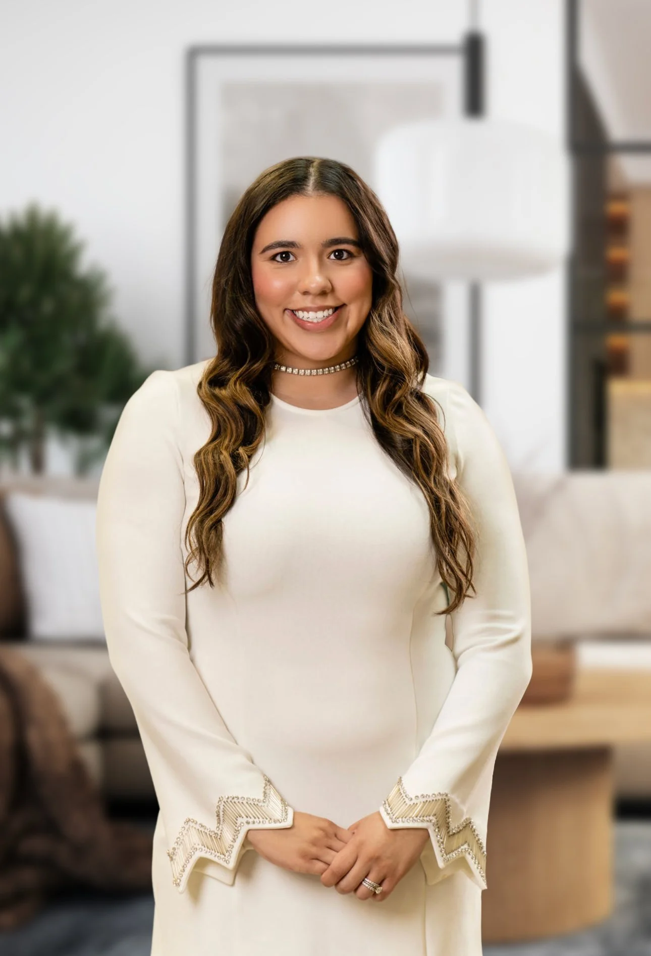A woman with long, wavy brown hair, smiling, wearing a white long-sleeved dress with decorative cuffs, and a pearl choker necklace, standing in a modern, well-lit living room with a Christmas tree in the background.