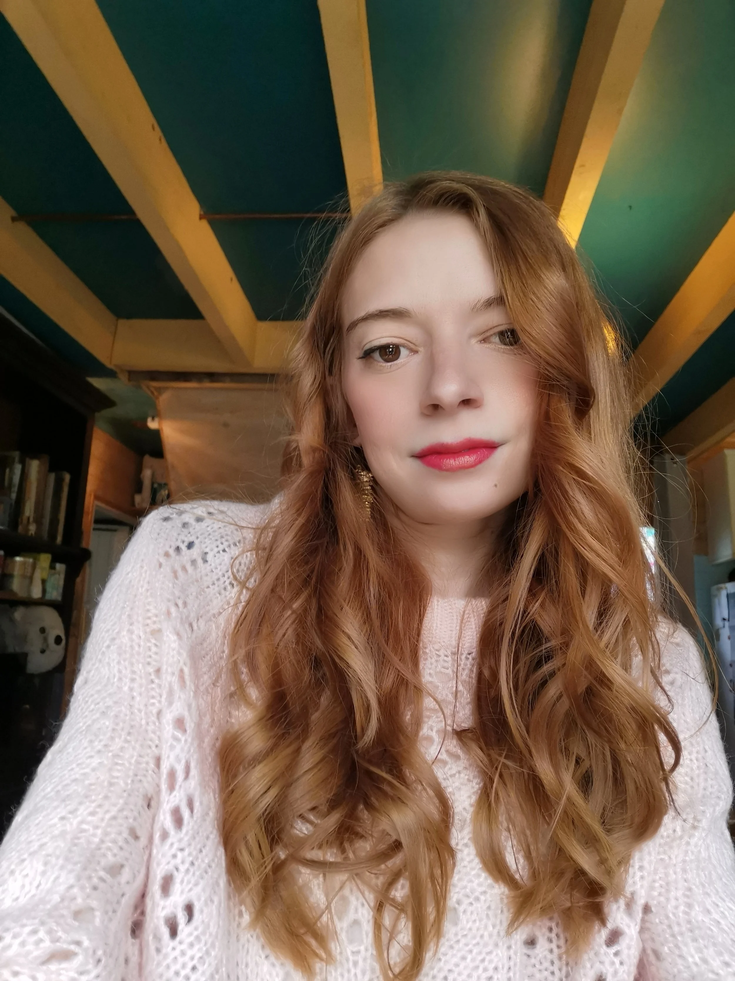 Jeune femme aux longs cheveux roux, portant un pull blanc, dans un intérieur avec plafond à poutres en bois peintes en vert et jaune.