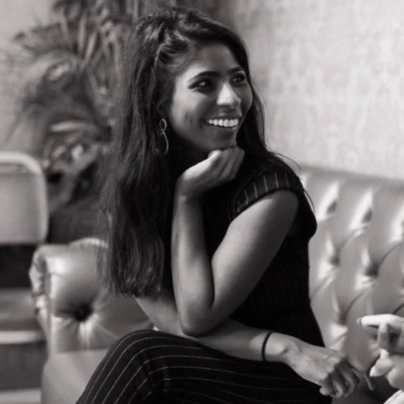 A young woman with long dark hair, smiling, sitting on a tufted leather sofa in a casual indoor setting.