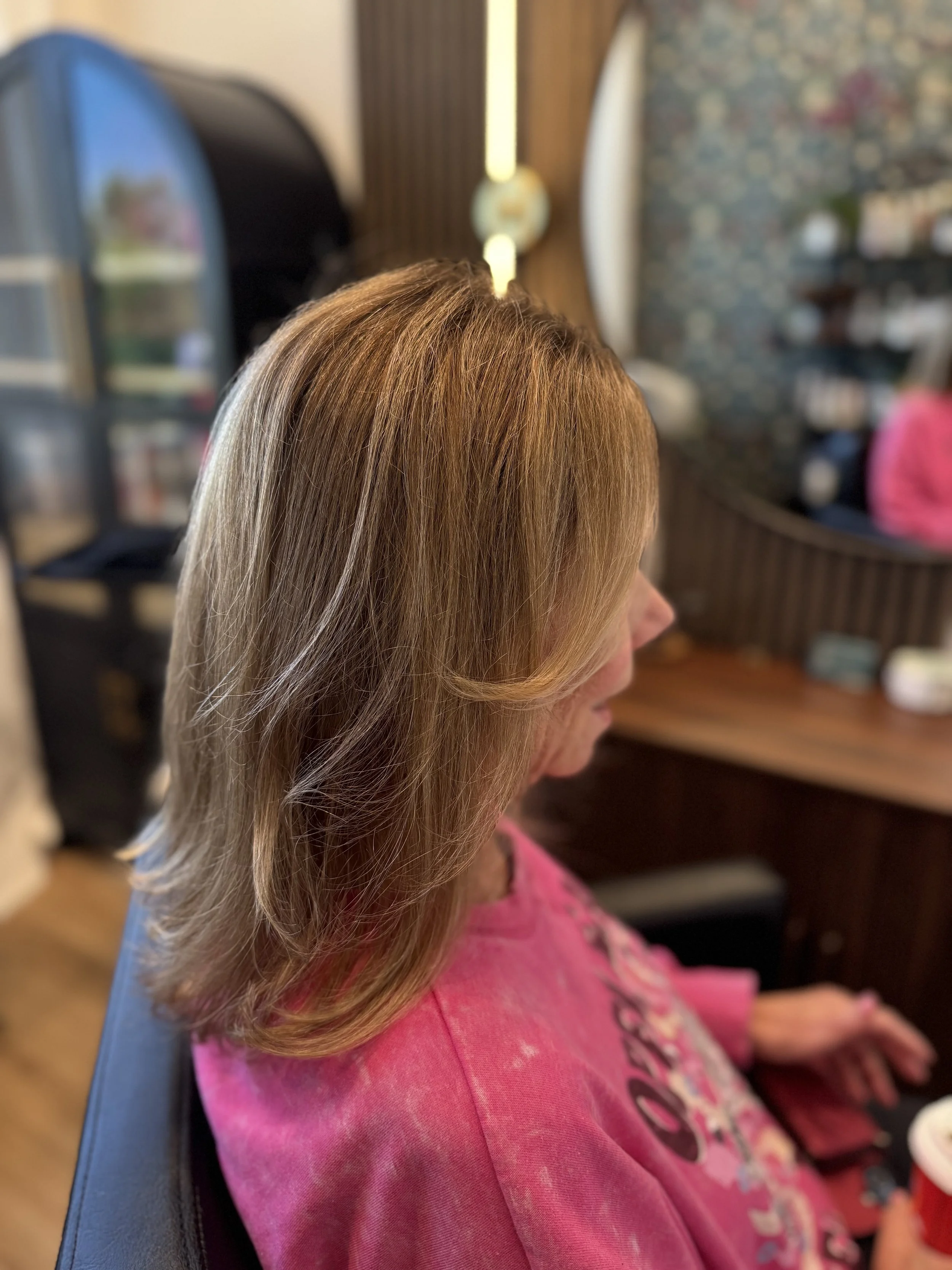 Side view of an elderly woman with shoulder-length light brown and gray hair, wearing a pink shirt with a floral design, sitting in a hairstylist's chair in a salon. Hair salon in Stony Plain, AB.