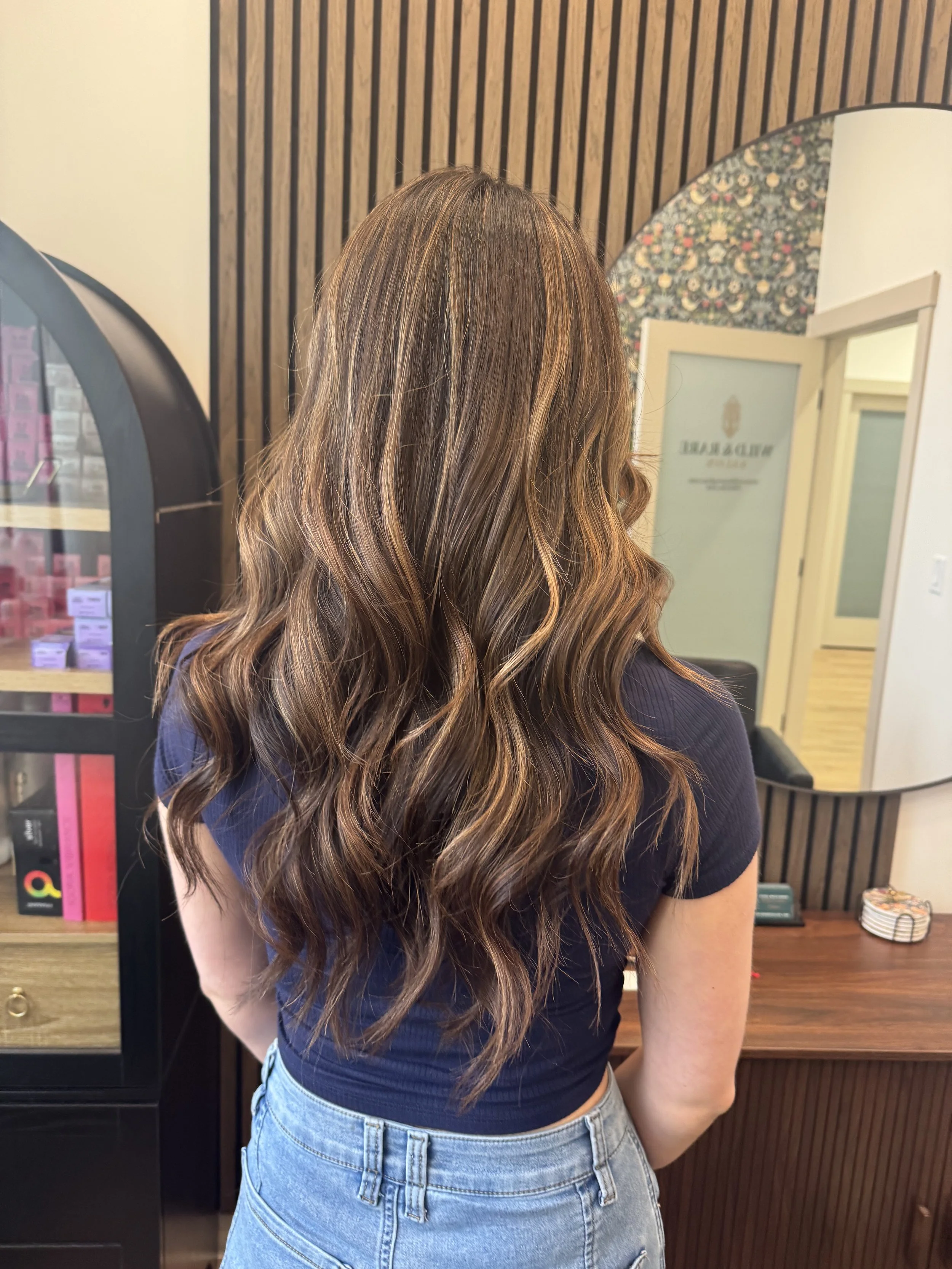 Back view of a woman with wavy, brown hair with blonde highlights, wearing a navy blue shirt and light blue denim jeans, standing in an indoor space with wooden wall panels and a mirror. Hair salon in Stony Plain, AB.