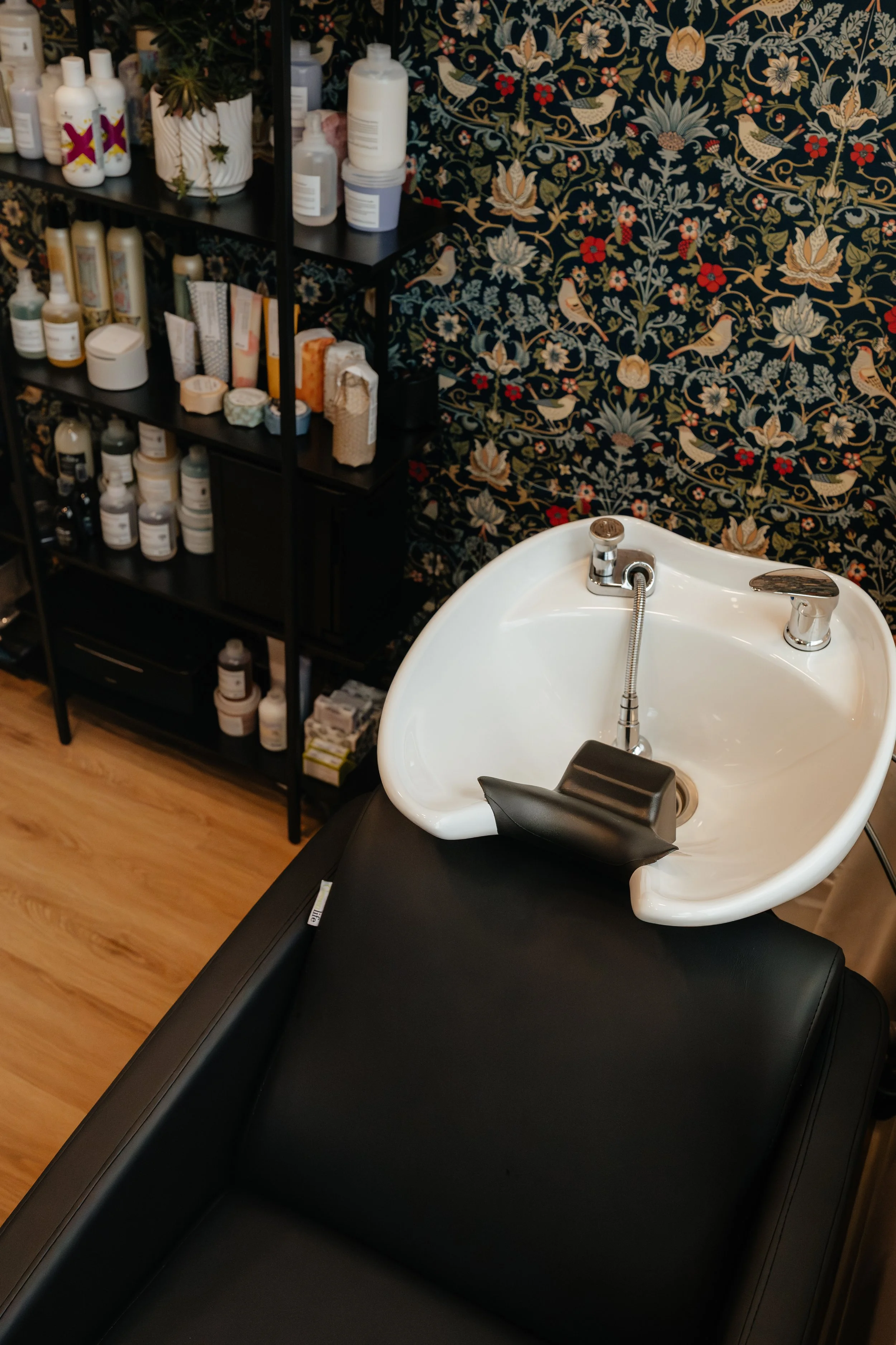 A black hair salon shampoo bowl with a headrest, next to black shelving filled with hair care products and supplies, against a floral-patterned dark wallpaper in Stony Plain, AB.