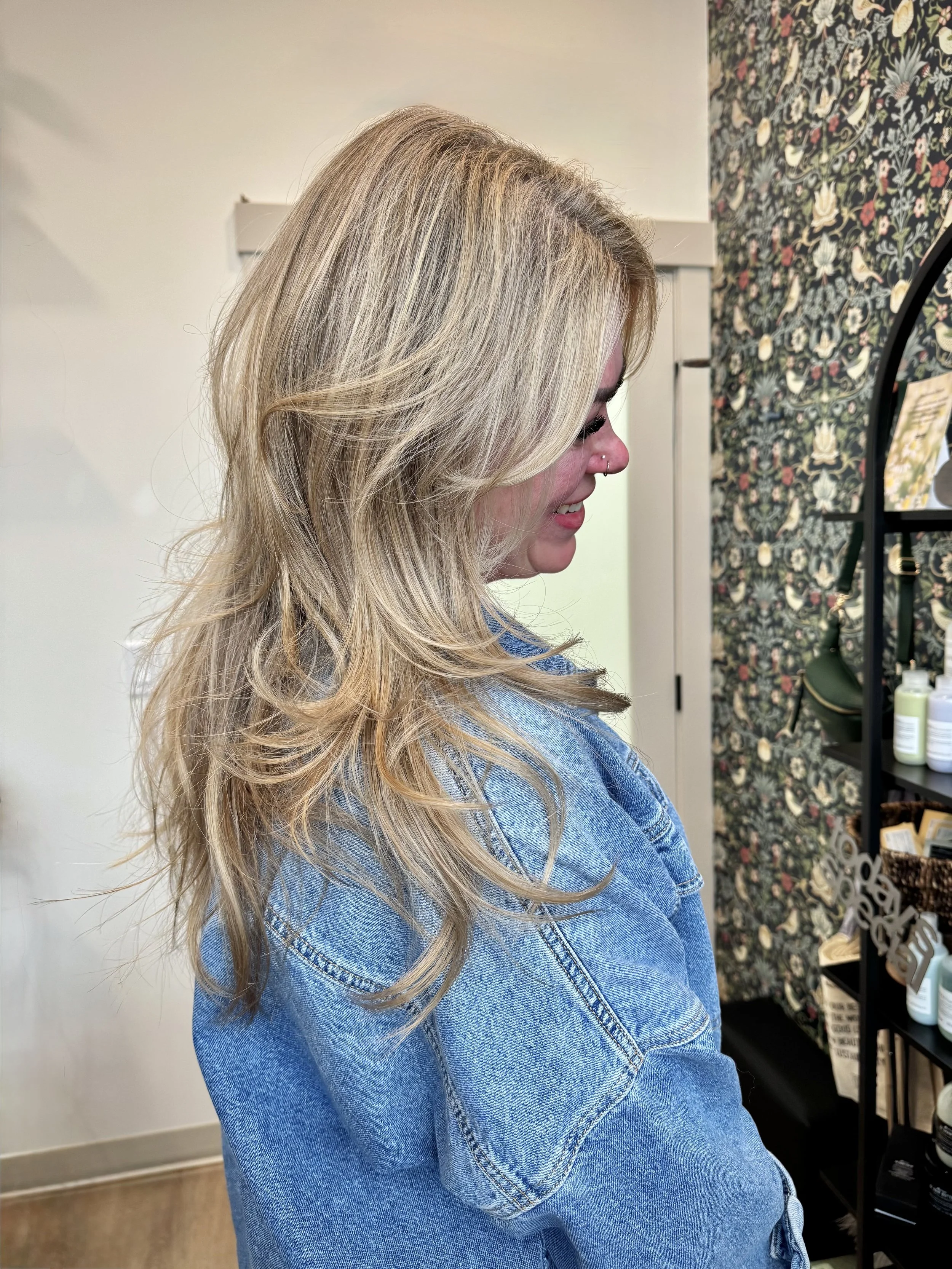 Side view of a woman with long blonde hair in a salon, wearing a denim jacket, smiling, with a floral wallpaper background and hair care products on a shelf. Hair salon in Stony Plain, AB.