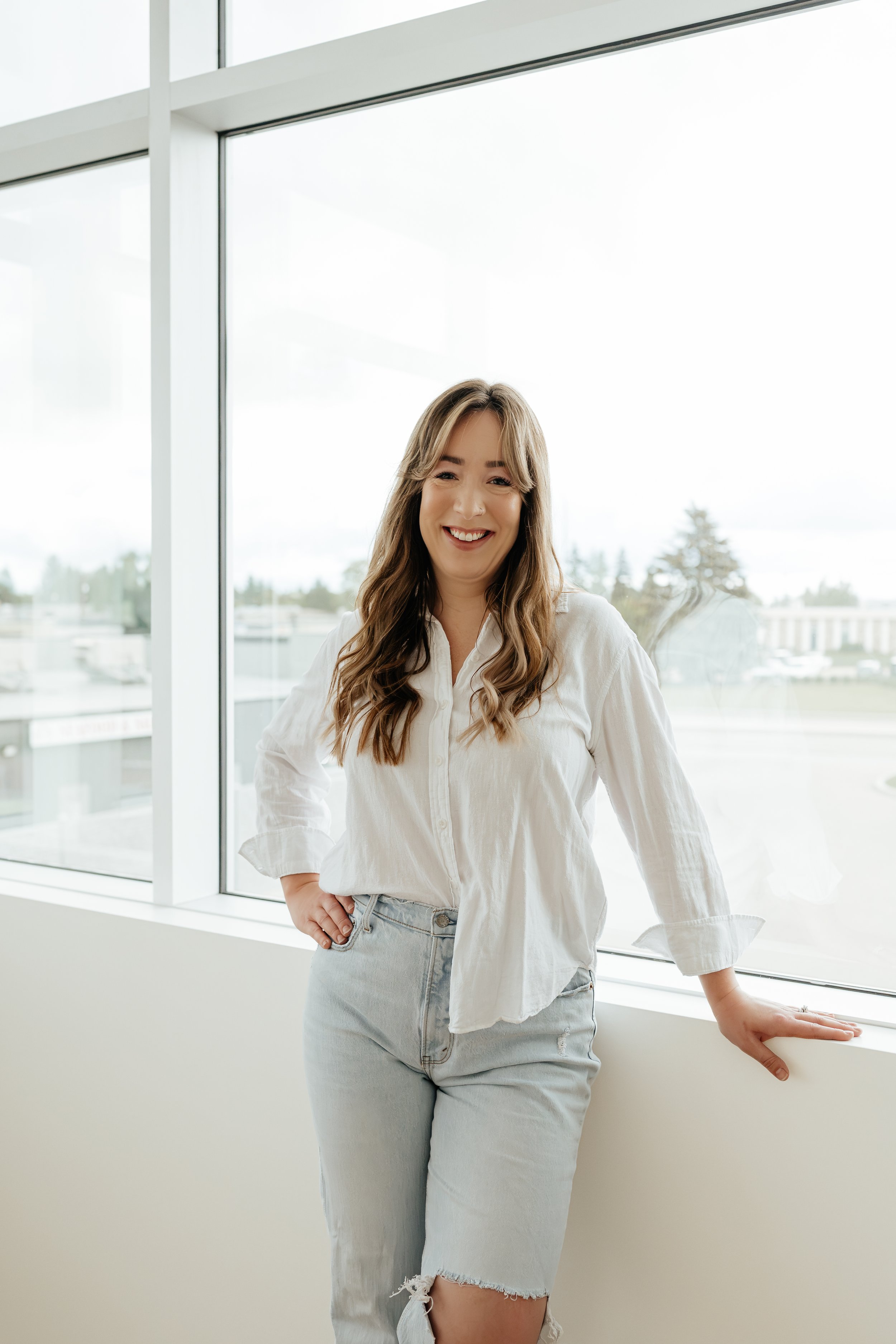 A woman standing by a large window, smiling and looking at the camera. She is wearing a white button-up shirt and light-wash jeans with a ripped knee, with her right hand resting on her hip and her left hand on the windowsill. The background shows an outdoor scene with trees and buildings.
