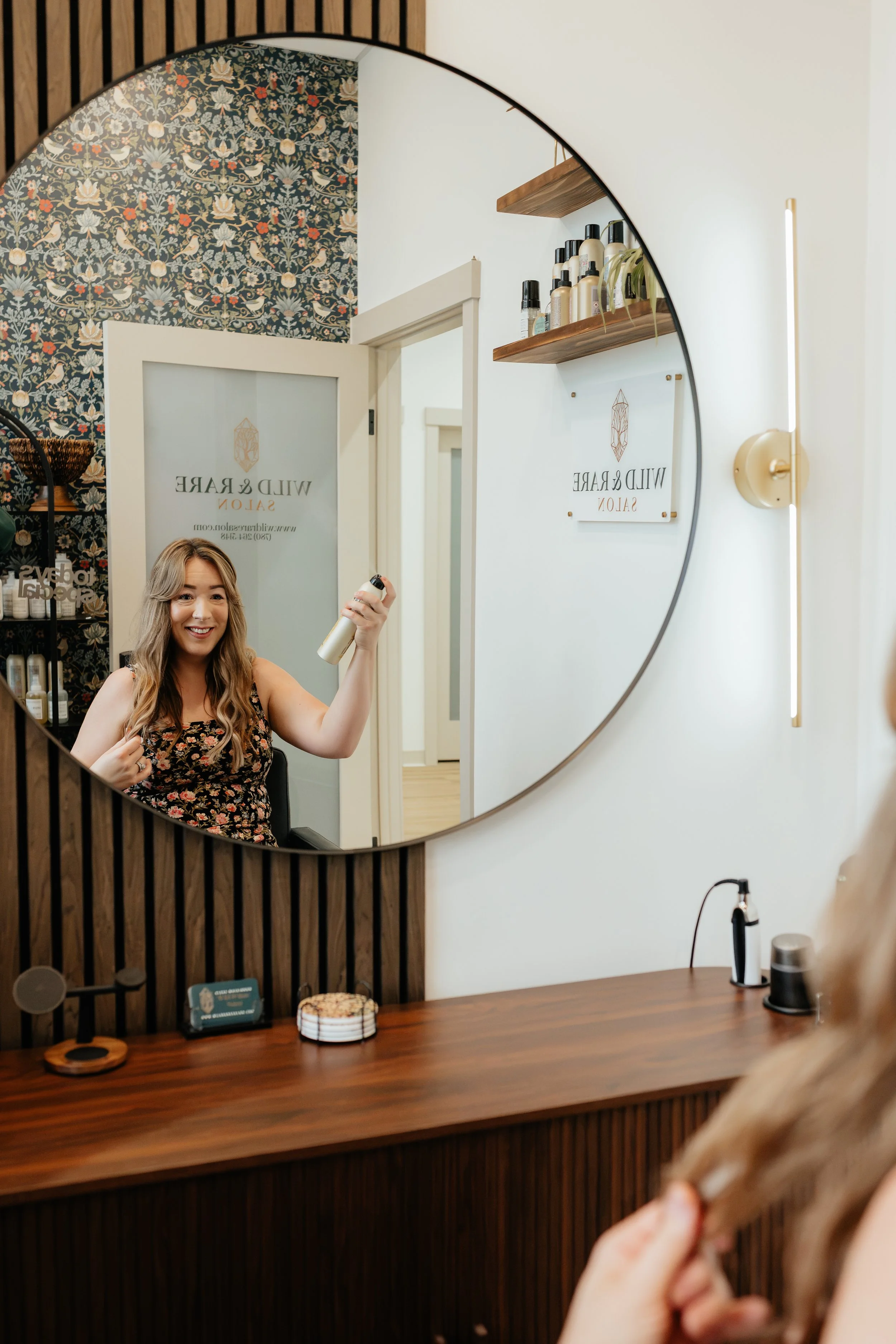 A hairstylist smiling in a hair salon in Stony Plain, AB holding a hairbrush, seen through a circular mirror that reflects shelves with hair care products and part of the salon's interior.