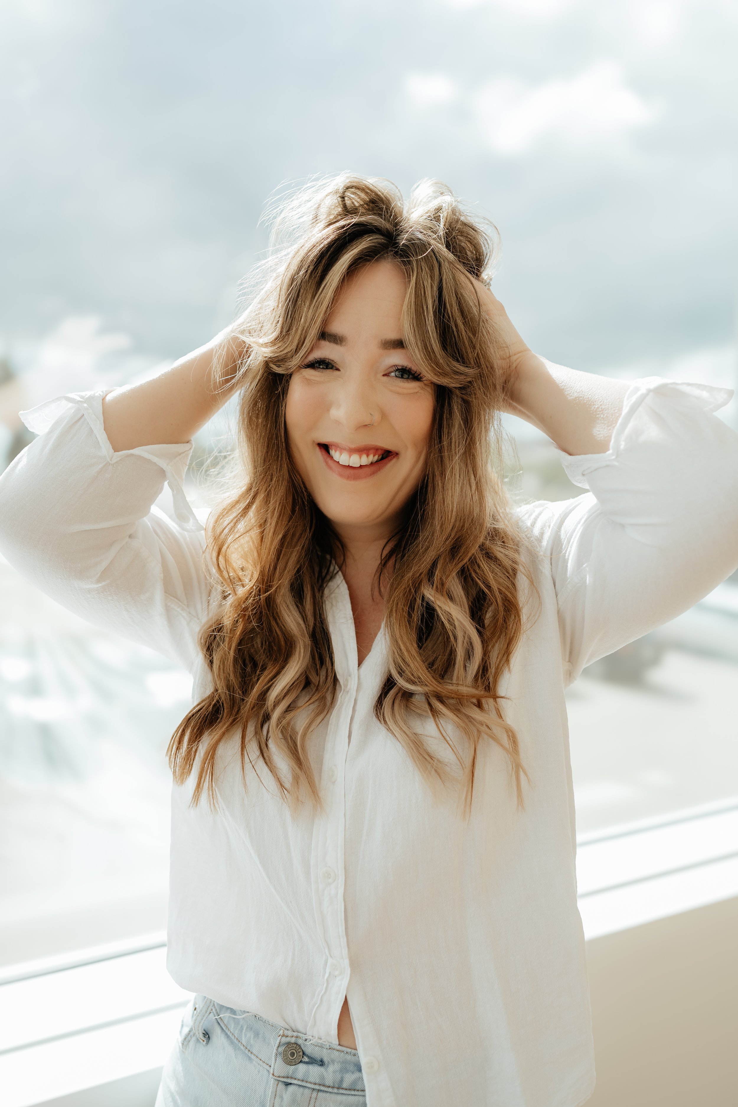 A young woman with long, wavy hair wearing a white button-up shirt and light blue jeans, smiling and holding her hair back while standing outdoors on a bright day with a cloudy sky in the background. Har salon in Stony Plain, AB.