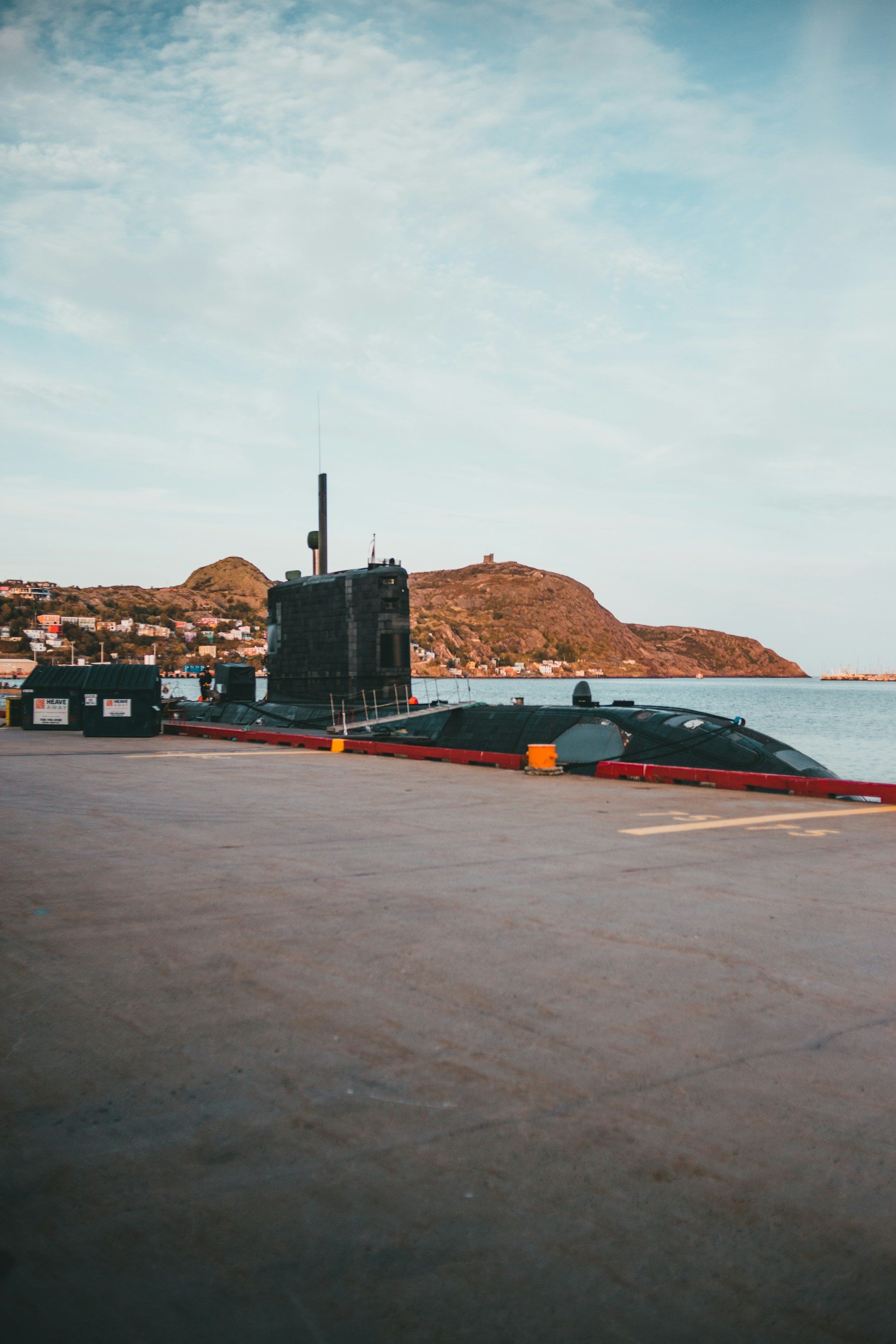A submerged submarine near a harbor with hills in the background and a partly cloudy sky.